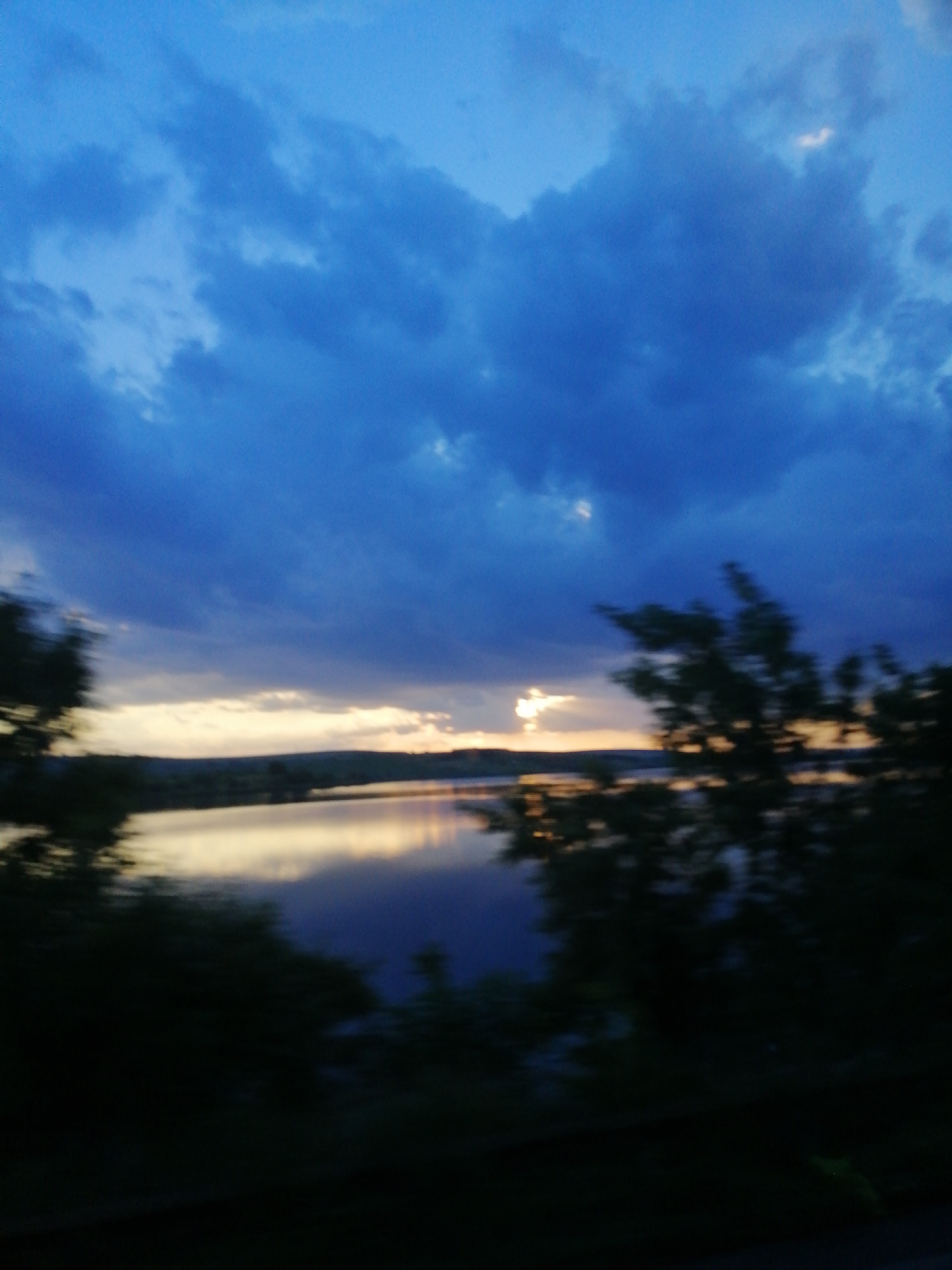 The image depicts a blurry, wide-angle view of a body of water at dusk or dawn. Dark, ominous clouds dominate the sky, with hints of yellow light breaking through near the horizon. Silhouetted trees and vegetation are visible in the foreground and along the shoreline, further obscuring details of the landscape. The water's surface reflects the muted colors of the sky, creating a somewhat tranquil yet melancholic atmosphere. The overall impression is one of a natural, undeveloped setting, possibly a lake or large river, under a dramatic sky.