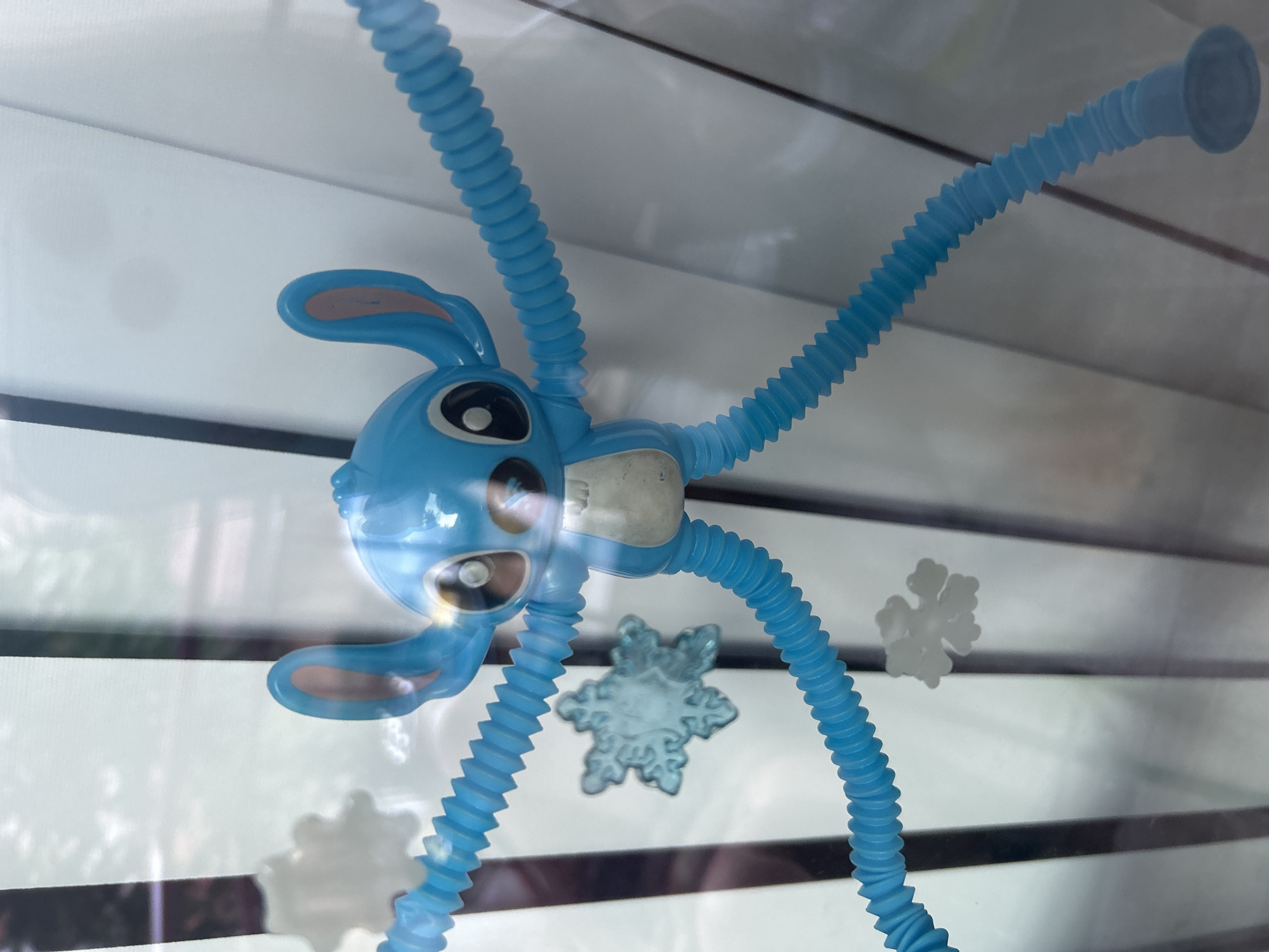 A blue toy resembling the character Stitch from Disney's Lilo & Stitch is positioned in the foreground, attached to a window. The toy has flexible, segmented blue arms and legs extending outwards. Two snowflake decorations are visible on the window behind the toy, one to the left and one to the right of its torso, appearing slightly out of focus. The background is dominated by the reflection of vertical dark lines and bright white areas, suggestive of a window frame and possibly blinds or curtains. The overall scene is viewed through glass, creating reflections and obscuring some details of the environment beyond. This image was captured on "Knyaz Alexander Battenberg" Street in Burgas, Bulgaria.