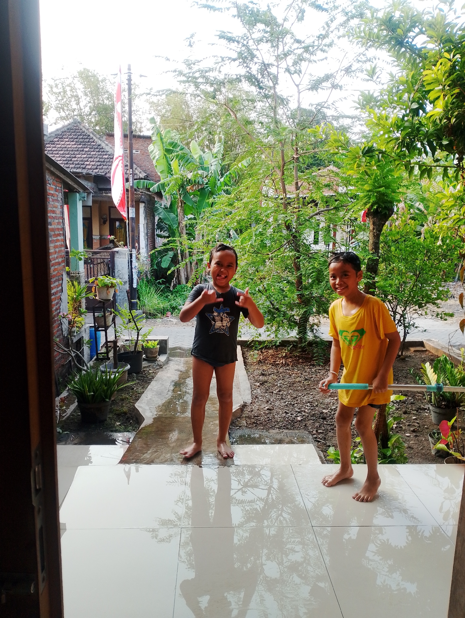 In the city of Plumbungan, Indonesia, two young boys stand on a wet tiled patio outside a house. The boy on the left, wearing a dark t-shirt and shorts, makes a rock-on hand gesture and sticks out his tongue. The boy on the right, in a yellow t-shirt and dark shorts, holds a blue and green water gun. Behind them, a red and white flag is attached to a pole, and lush green trees and plants surround the area, with parts of a brick house visible. The ground is scattered with dirt and pebbles, and the tiled floor is wet, reflecting the trees and the sky.