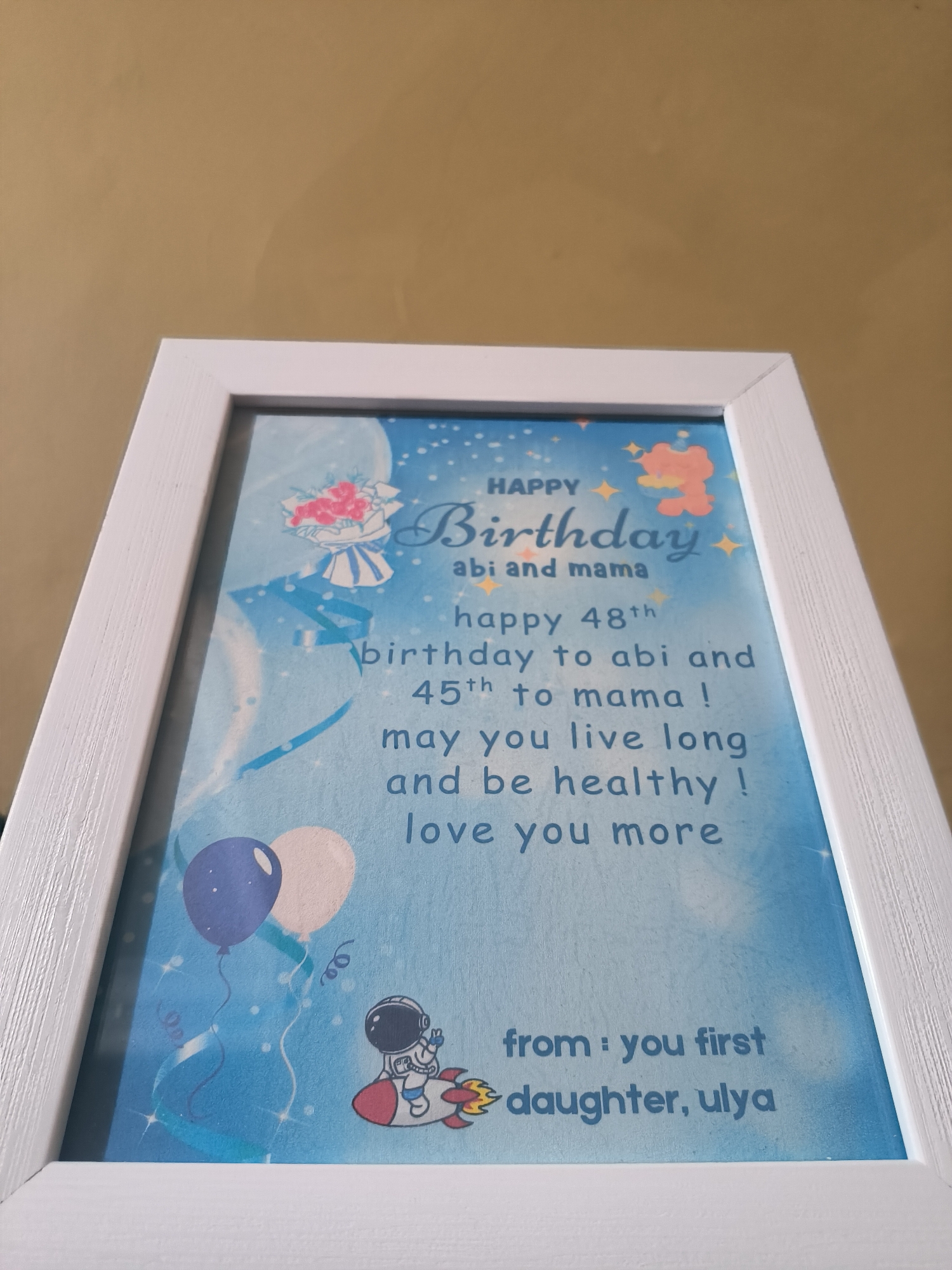 A framed birthday card is shown against a neutral, solid-colored wall. The card has a white frame and a blue background. The text on the card reads "HAPPY Birthday abi and mama happy 48th birthday to abi and 45th to mama! may you live long and be healthy! love you more from: you first daughter, ulya." The card also features illustrations, including a bouquet of flowers, a teddy bear, balloons, and an astronaut riding a rocket. The location, Lhokseumawe, Indonesia, is not discernible in the image.