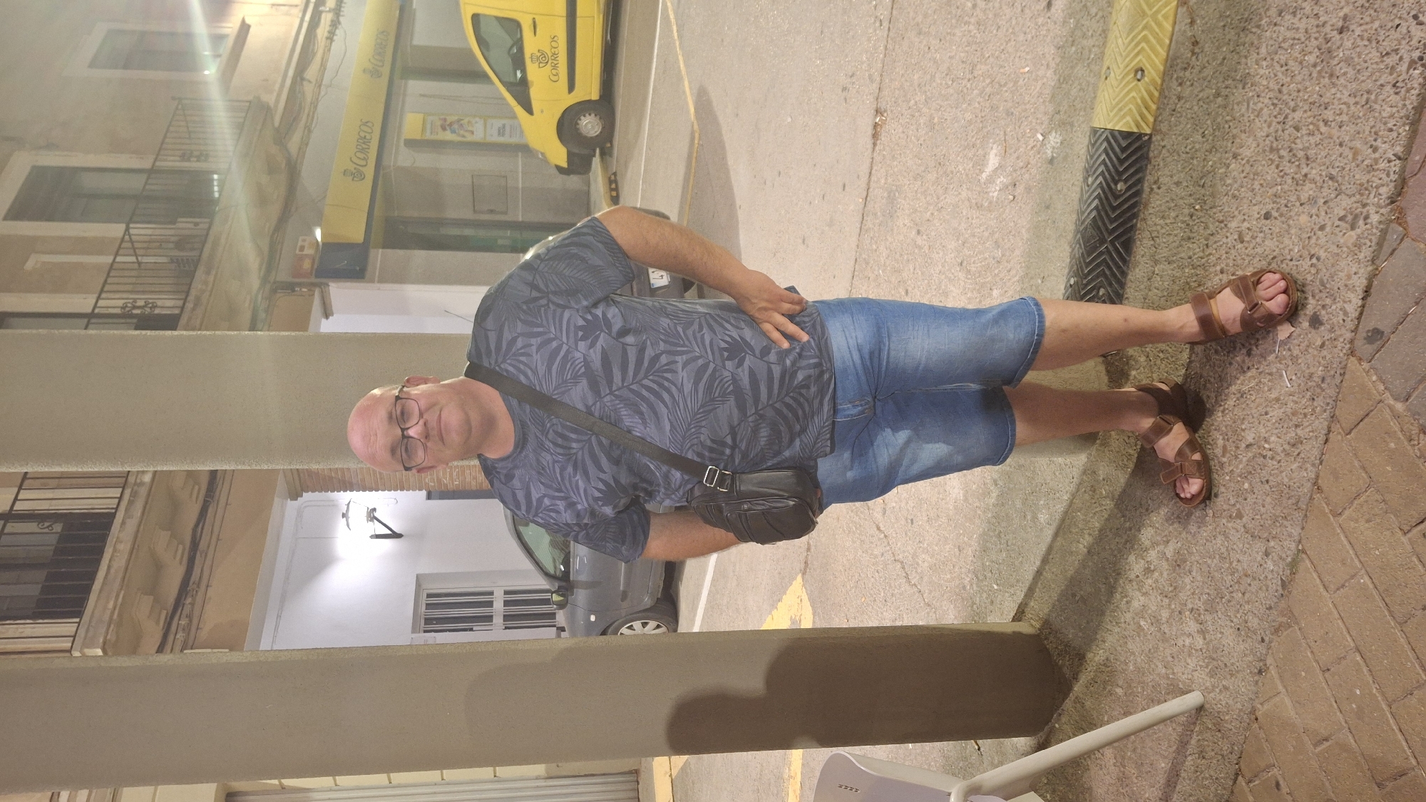 A bald, fair-skinned man wearing glasses, a dark t-shirt with a leaf pattern, denim shorts, and brown sandals stands in front of a building with a yellow "Correos" (Spanish postal service) sign. He has a black shoulder bag. A gray car and a yellow Correos van are visible in the background. The setting appears to be a street or plaza in Híjar, Spain, with cobblestone pavement in the foreground and a concrete sidewalk. The lighting suggests it is evening or nighttime.
