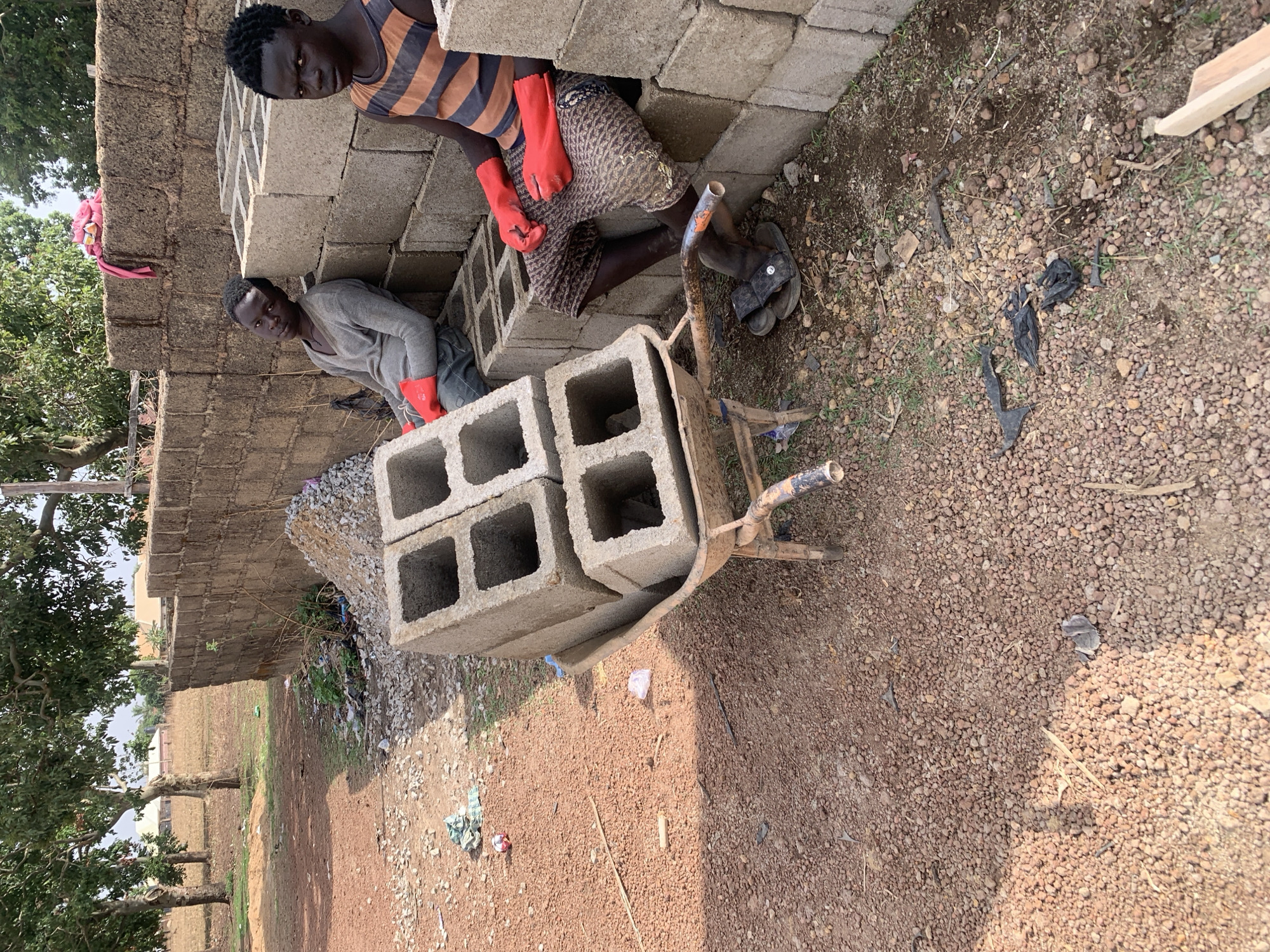In Rafin Bauna, Nigeria, two individuals are engaged in construction-related activities. One person, wearing a striped tank top and red gloves, is positioned near a stack of concrete blocks. The other person, wearing a gray shirt and red gloves, is next to a wheelbarrow loaded with more concrete blocks. The wheelbarrow is resting on a gravelly ground. A brick wall stands in the background, with trees and a field visible beyond.