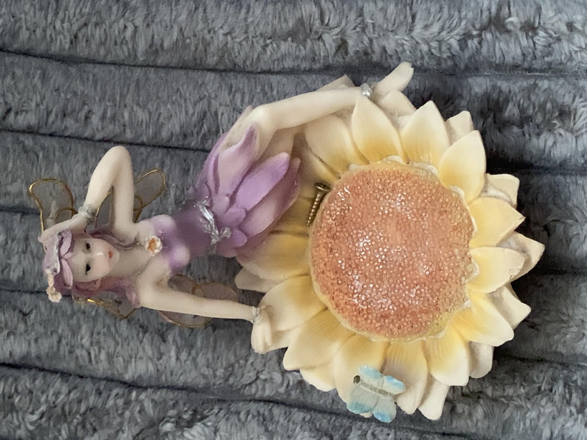 A figurine of a fairy with purple hair and a lavender dress sits on a large, yellow sunflower. A small, blue butterfly is perched on one of the sunflower petals. The figurine is positioned on a textured gray surface, possibly a blanket or rug. The background is out of focus. There is no discernible location in Tashkent, Uzbekistan, or any other city.