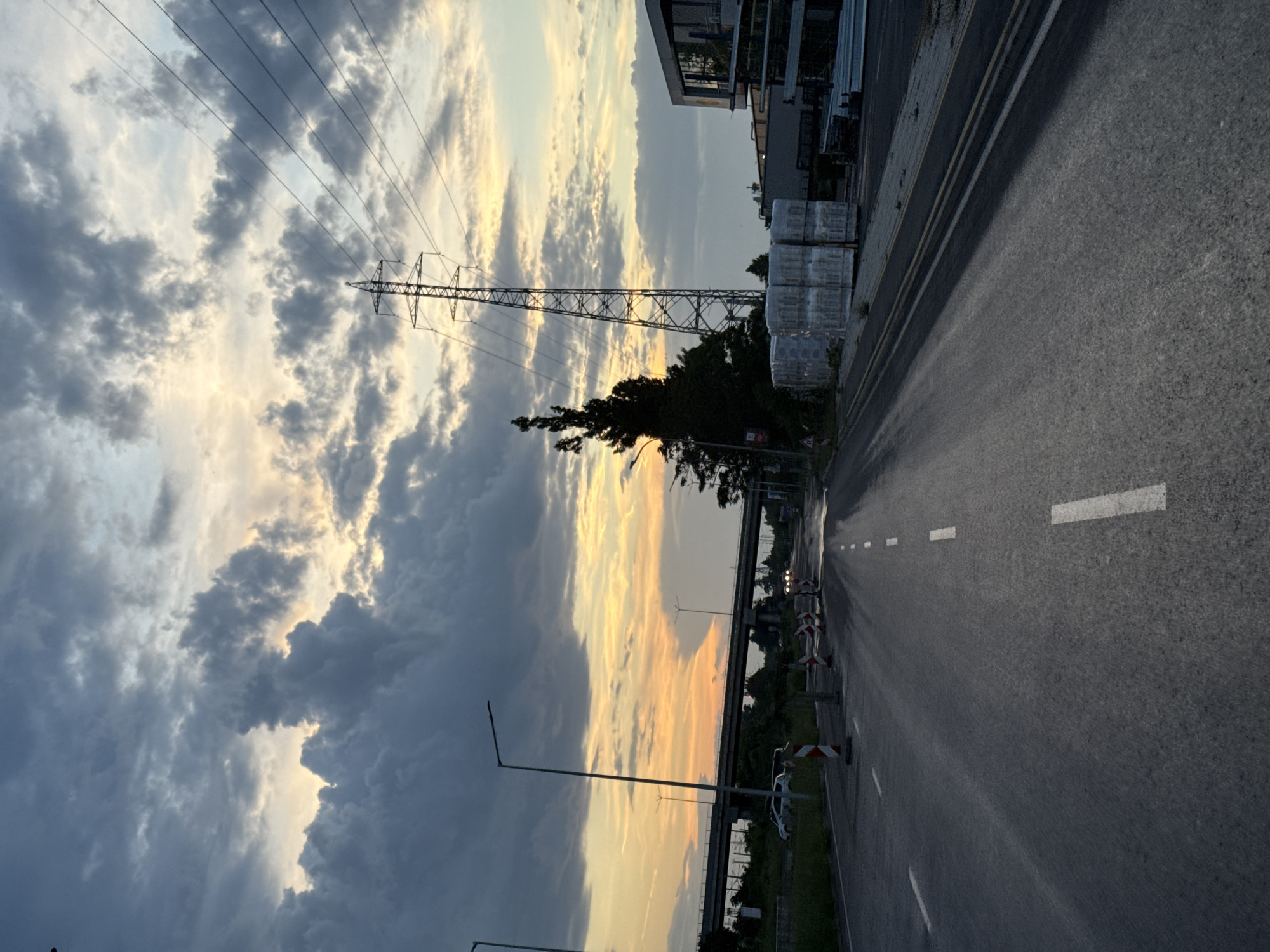 The image shows a road with white lane markings, a gray asphalt surface, and various road construction warning signs along the side. A power line tower is visible in the background, set against a cloudy sky with streaks of sunset colors. Adjacent to the road is a building with stacked, shrink-wrapped pallets. The location appears to be in an industrial or commercial area of Burgas, Bulgaria, with a bridge visible in the distance. The scene is captured at dusk or dawn, with the lighting suggesting a transitional time of day.