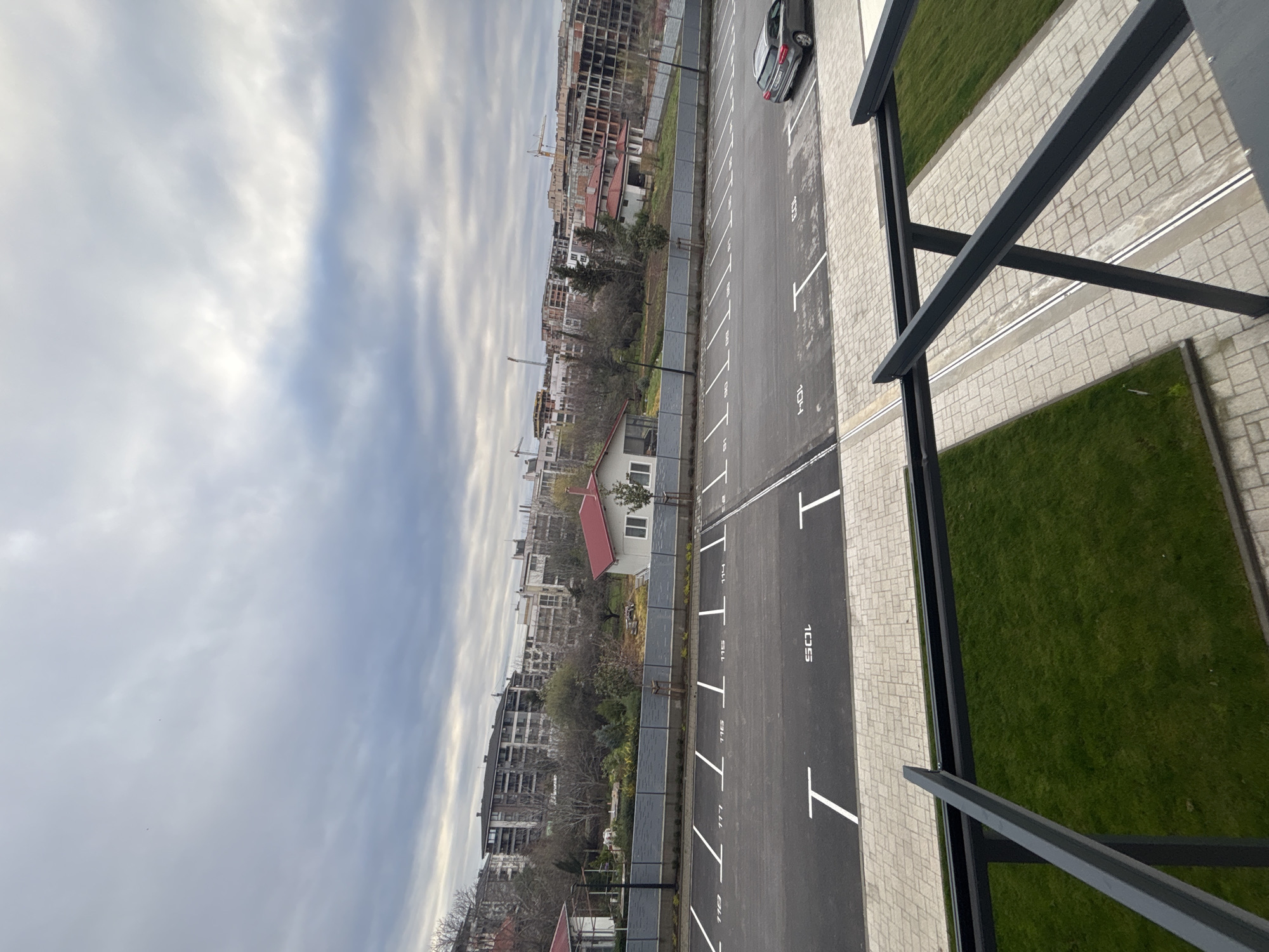 This image shows an outdoor parking lot next to a building under construction. The parking lot has numbered parking spaces marked in white. Adjacent to the parking lot is a landscaped area with grass. In the background, buildings under construction and residential buildings are visible. The sky is overcast. There is no visible evidence to confirm that this image is in the city of Plovdiv, Bulgaria.