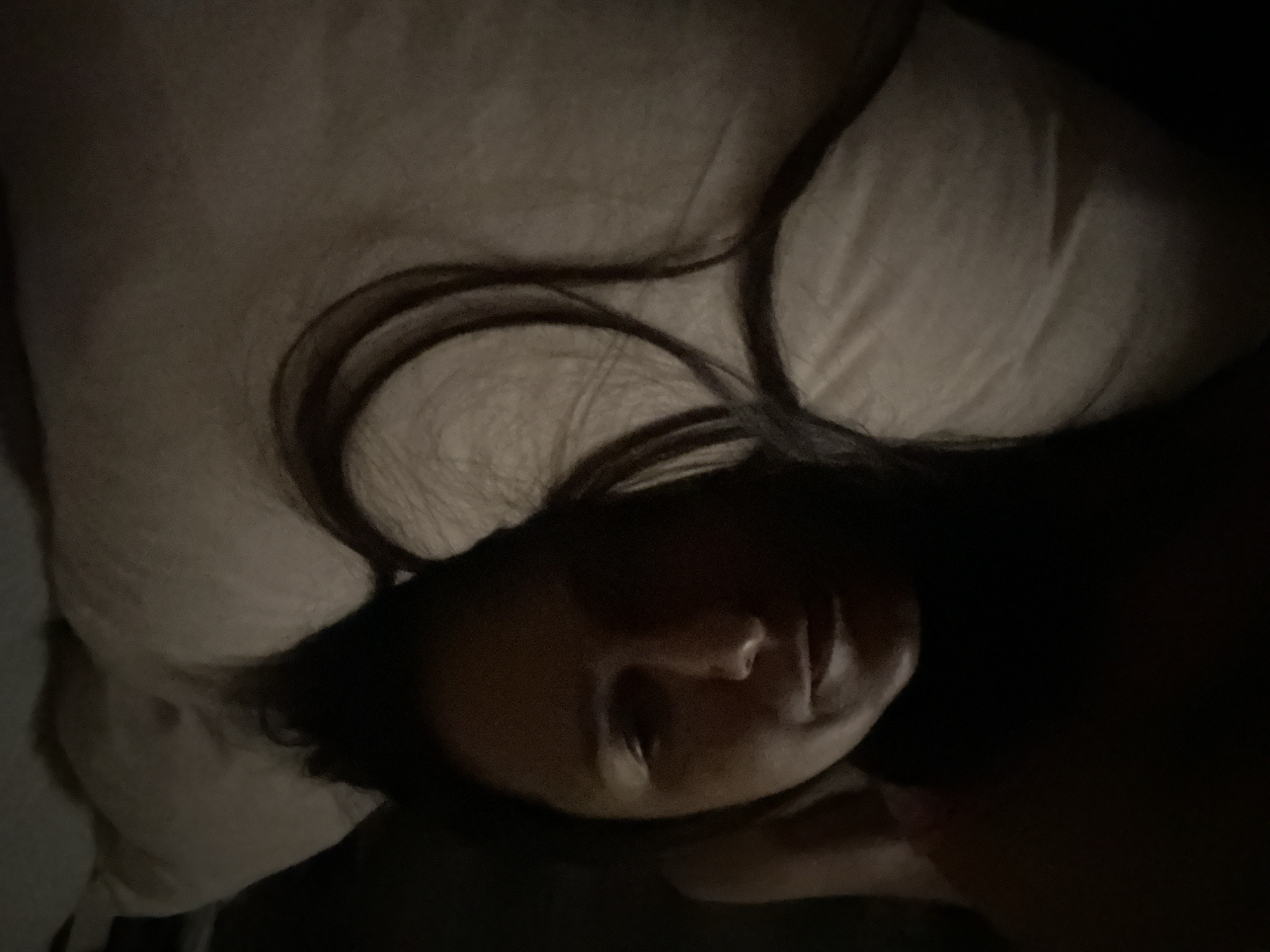 A person with long, dark hair is lying on a pillow. The person's face is partially illuminated, revealing their nose and mouth. The background is dark, and the pillow appears to be light-colored. There is no indication of the location being in London, United Kingdom.