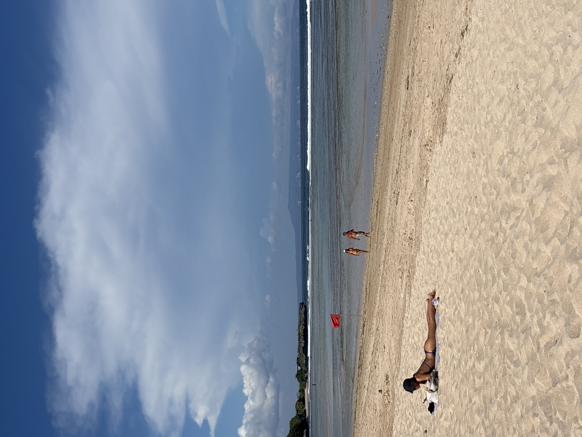 The photograph shows a beach in Nusa Dua, Indonesia. The sky is blue with white clouds. A calm, shallow ocean meets the sandy beach. Two people are wading in the water near a red flag planted in the sand. Closer to the foreground, a person is lying on a white towel on the beach.
