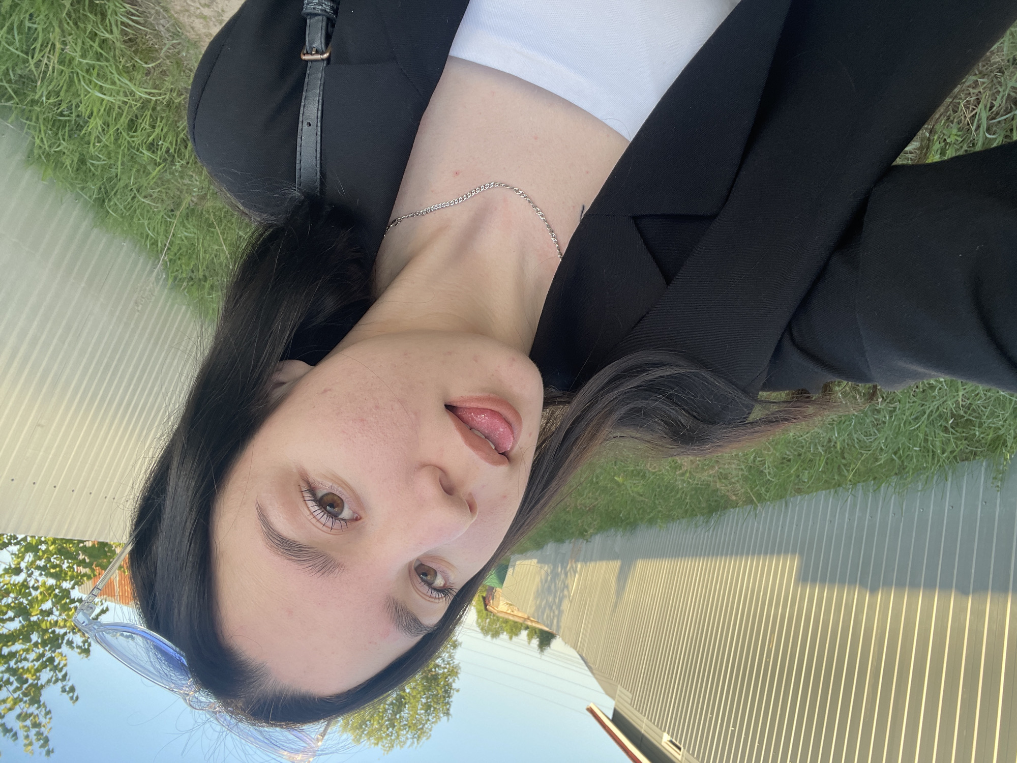 A young woman is posing outdoors in Ольховка, Russia. She has dark hair and brown eyes, and is wearing a white top and a black blazer. A silver necklace is visible around her neck. Her tongue is slightly sticking out. Sunglasses are positioned on top of her head. Behind her, there is grass, a metal fence, and a glimpse of trees and sky.