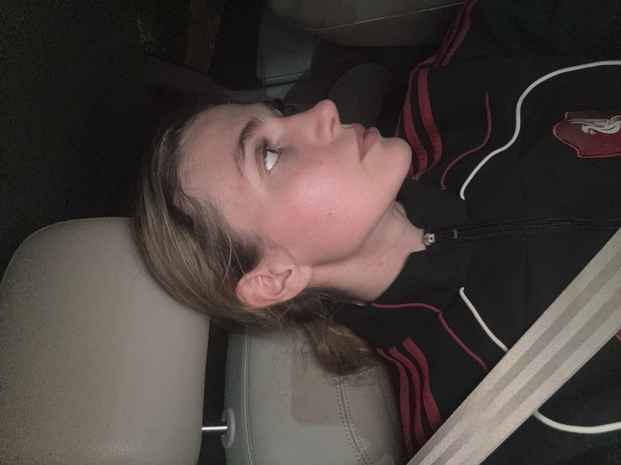 The image captures a young person, likely female, resting in a car seat. The subject is seen in profile, with their head turned towards the right and looking upwards, suggesting a gaze towards the car's roof or a window. Their eyes are open, and their expression appears calm or contemplative. The person's face shows a natural rosy complexion.
They are wearing a black zip-up jacket with distinct red stripes running along the shoulders/sleeves and a thin white stripe or piping on the front. On the right side of the jacket's chest (viewer's left), there is a red and white emblem or patch. While the details are not entirely clear, it seems to depict an open book or a crest-like design, but no legible text is visible. A light-colored seatbelt extends diagonally across their chest.
The person has light brown or blonde hair, which appears slightly disheveled from resting. They are leaning against a light beige or grey car headrest. The lighting in the image is dim, indicating it might be nighttime or a poorly lit interior. The overall scene suggests a quiet moment during a car journey. While the location context is Tbilisi, Georgia, no specific visual cues within the image confirm this.