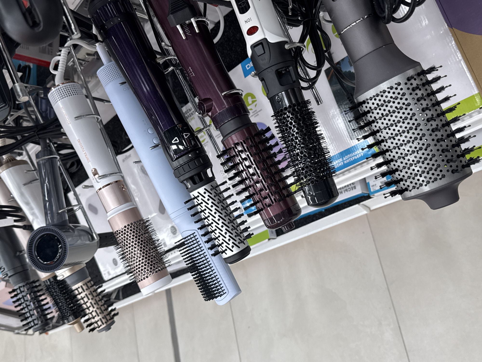 The image shows a retail display of various hair styling tools, specifically ionic hair brushes and dryers. The items are arranged on a tiered metal rack, with several of them hanging from hooks. The brushes vary in size, color, and bristle type, with some featuring black bristles and others a mix of black and metallic. The handles are in shades of gray, blue, purple, and white, with some displaying brand names and power ratings like "1000" and "ION". The background consists of a light-colored, tiled floor and hints of product packaging. The context suggests a store environment, likely in the city of Mikhaylovsk, Russia, given the presence of Cyrillic text on some of the packaging and product labels, such as "ВЕСЫ НА" which likely translates to "Scales ON" or similar phrasing relating to weight or balance. No people are visible in the image.