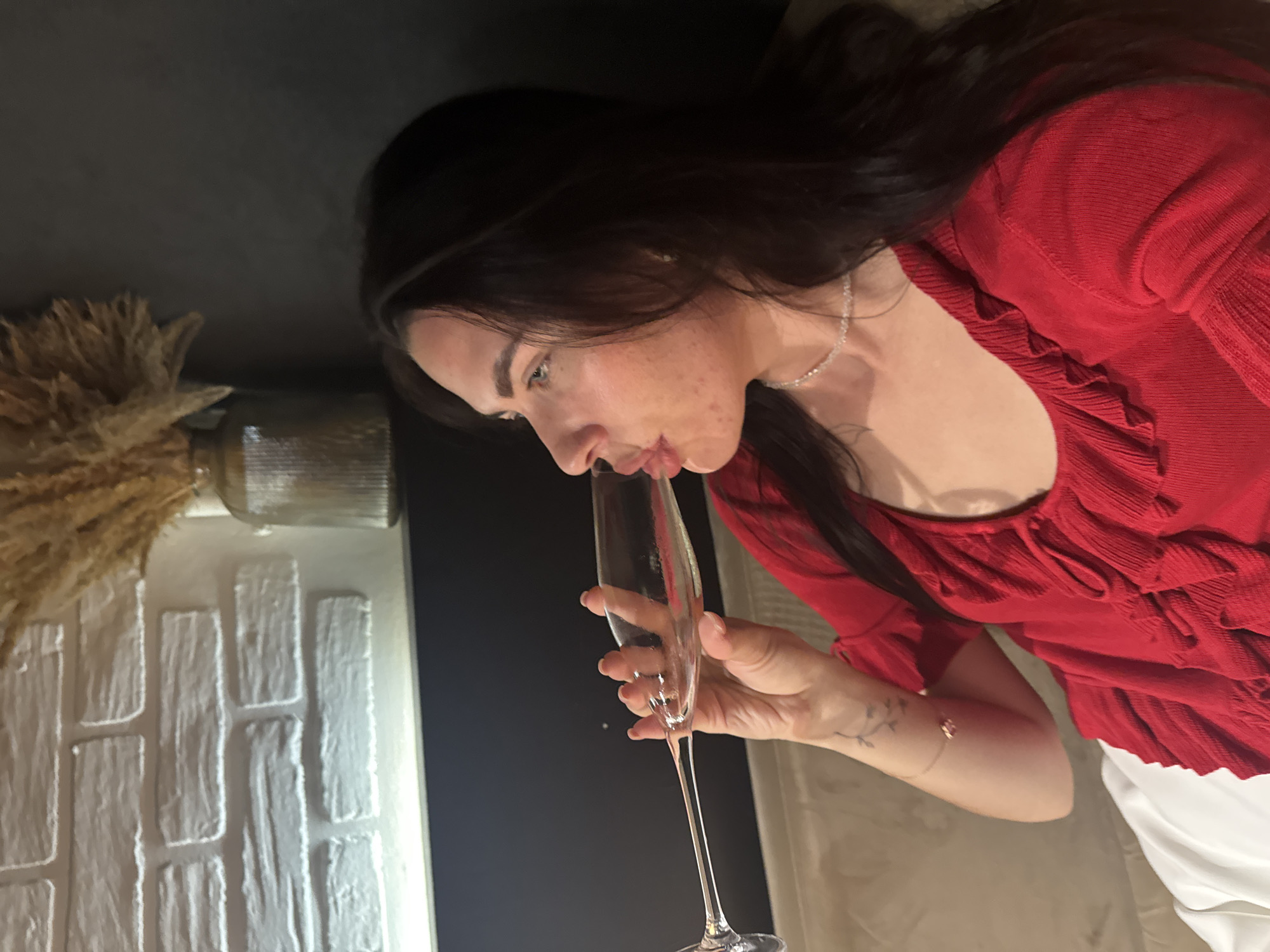 A woman with dark hair and fair skin is captured in a side profile, wearing a red ruffled top and a delicate necklace. She is holding a champagne flute to her lips, as if drinking. Her left arm, adorned with a tattoo and a bracelet, is visible. The background features a white brick wall with ambient lighting and a vase containing dried wheat-like stalks. The scene appears to be indoors, possibly in a domestic setting or a restaurant, with a dark wall and a light-colored couch or seating area. No specific location in Chaikovsky, Russia is discernible from the image.
