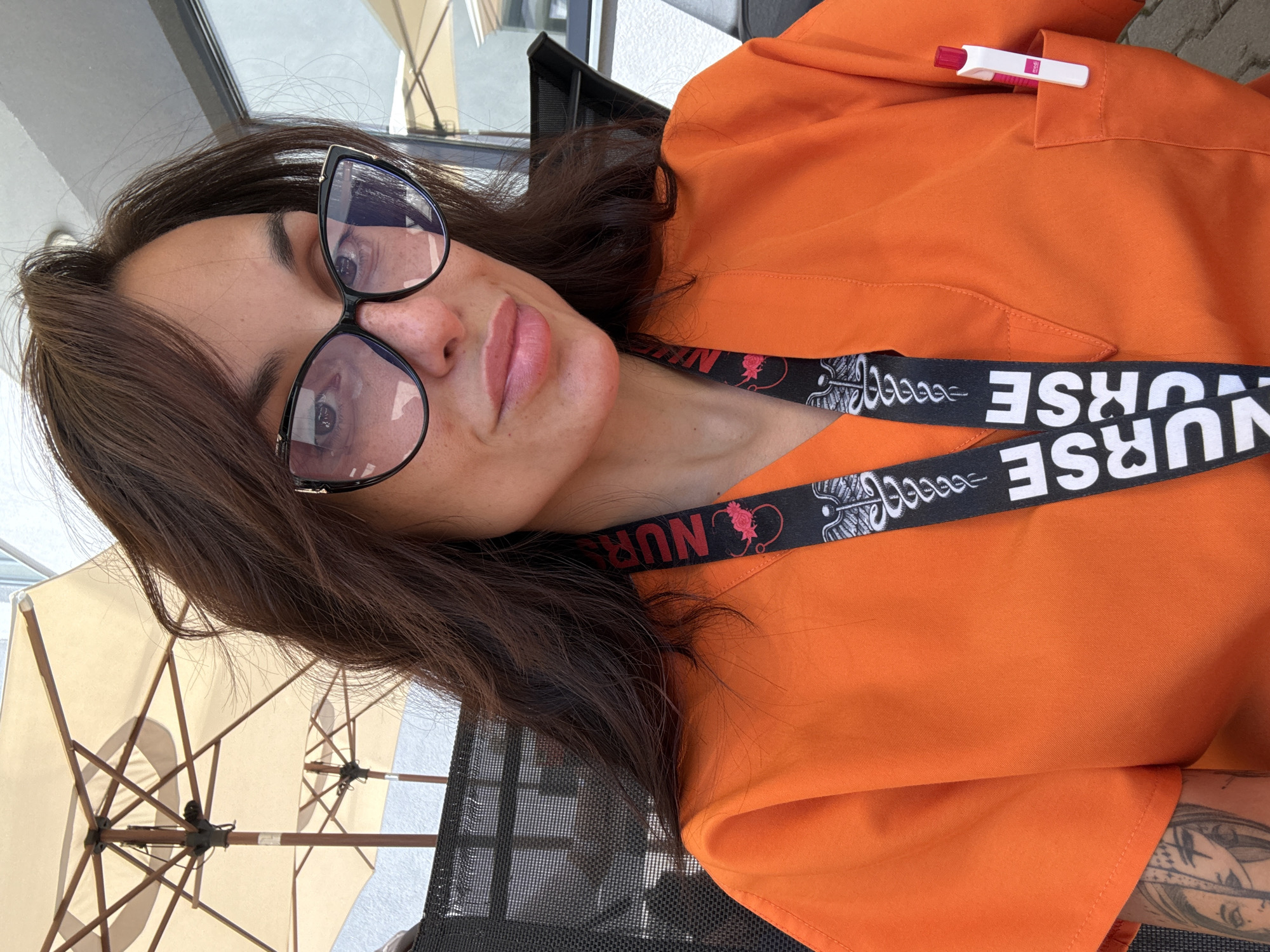 A woman is pictured from the chest up, wearing an orange scrub top and a lanyard that says "NURSE" in bold white letters. She has dark brown hair and is wearing large black-framed glasses. She is also wearing makeup and has freckles on her nose and cheeks. A pen is visible in the pocket of her scrub top. In the background, there are two outdoor umbrellas and a building with windows. The scene appears to be outdoors, possibly on a patio or balcony, with natural light. The setting is consistent with a cafe or an outdoor seating area in Pleven, Bulgaria, given the context.