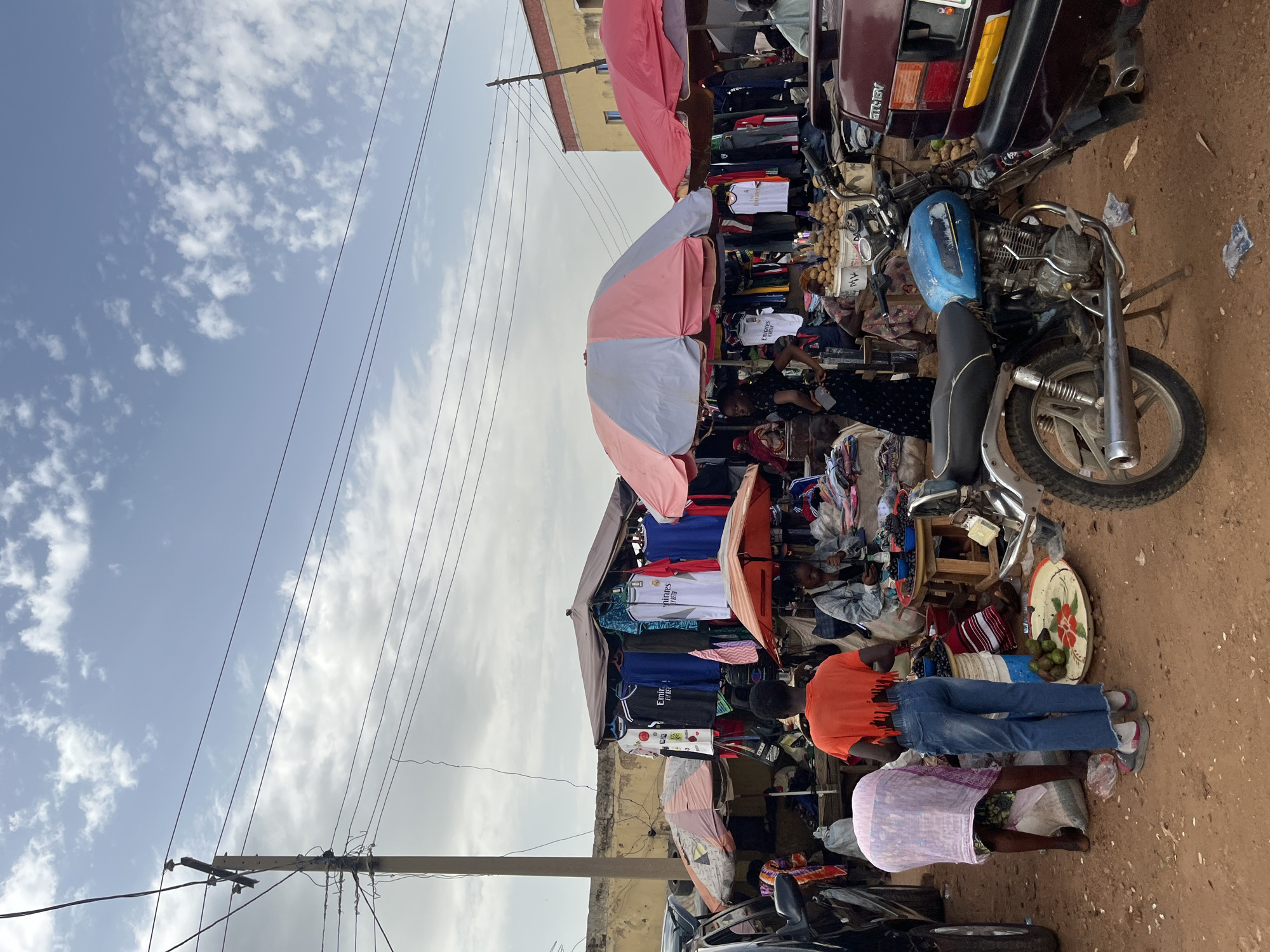 The image depicts an outdoor market scene in Rafin Par, Nigeria, under a partly cloudy sky. The market appears to be selling clothing, with numerous shirts hanging on display under tarps and awnings. Several people are present, engaged in shopping or manning the stalls. A motorcycle and a maroon car are parked within the market area, suggesting transportation is a key element of the setting.
In the foreground, a person wearing an orange t-shirt and blue jeans stands next to another person wearing a purple garment. They are positioned near a display of goods, possibly for sale. Further back, more clothing racks are visible, showcasing a variety of t-shirts, some with visible text such as "Emirates Fly Better". The overall atmosphere seems busy, typical of a local market.
The ground is covered in a reddish-brown dirt, common in many regions. Electrical wires are strung overhead, indicating the presence of infrastructure. The time of day is difficult to determine precisely, but the lighting suggests it is daytime, possibly late morning or afternoon, given the bright sky. The weather appears to be warm, with the clouds suggesting a break from direct, intense sun. The market is set up along a street or open area, with some buildings visible in the background. The presence of the car and motorcycle, along with the variety of goods, points to a lively commercial hub within the community.