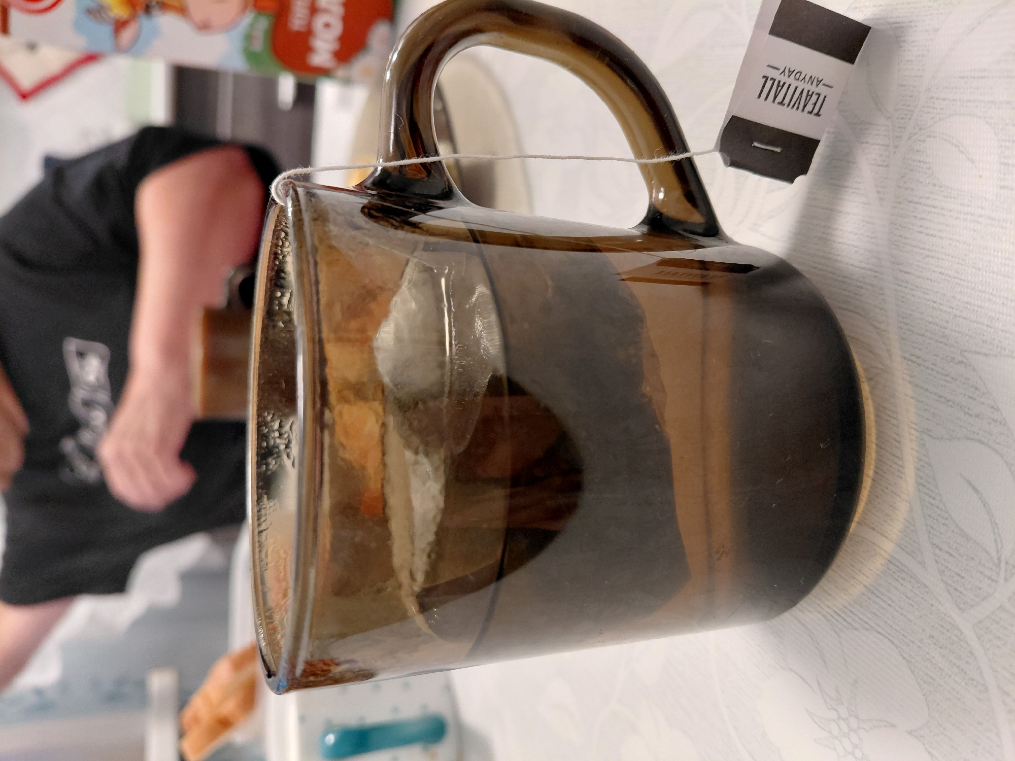 A close-up, angled shot shows a brown glass mug with a tag attached to its handle by white string. The tag is black and white with the text "TEAVITALL - ANYDAY" printed on it. The mug appears to be resting on a white surface with a floral pattern. In the blurred background, a person wearing a black t-shirt is visible, with their arm and shoulder in focus. The scene is indoors, likely in a domestic setting, with soft lighting. There are no obvious cues for time of day or weather.