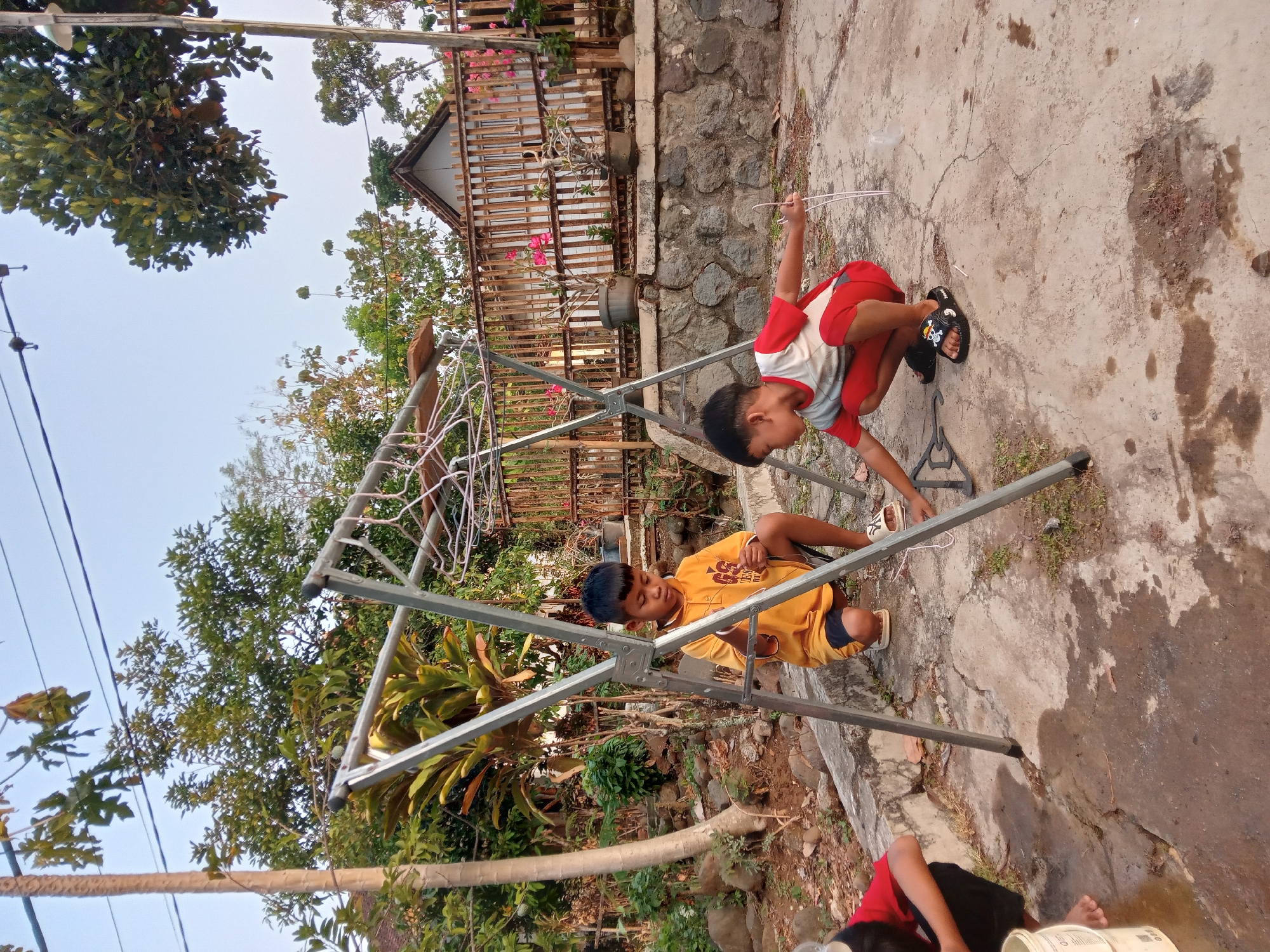 This is an outdoor photograph taken in Kedunggudel, Indonesia, showing three children interacting with a metal drying rack. The scene is set on a concrete patio with a stone wall and wooden fence in the background.
In the foreground, two boys are actively engaged. The boy on the left, wearing a yellow t-shirt and dark shorts, is crouched next to the drying rack, appearing to be holding something thin and white. The boy on the right, dressed in a red and white t-shirt and red shorts, is also crouched and holding two thin white strands, possibly strings or wires. His right hand is extended towards the drying rack. A third child, partially visible on the far left, is wearing a red shirt and black shorts and is crouched near a white bucket, appearing to be washing something.
The drying rack is a prominent feature, made of metal tubing with several hangers attached to its upper bars. The ground is a mix of concrete and dirt, with some scattered rocks and dried leaves. Vegetation, including trees and plants, is visible throughout the background, with a house and a wooden fence visible behind the stone wall. The sky is clear and bright, suggesting daylight. The overall atmosphere is casual and indicative of children playing outdoors.