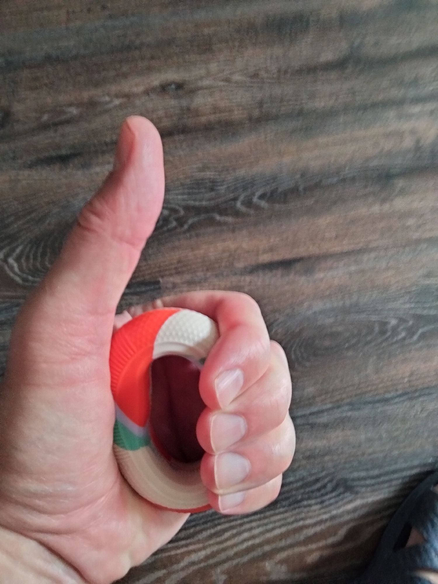 A hand holds a colorful ring-shaped stress ball against a wooden floor. The stress ball has an orange outer ring, a white textured middle section, and a green and white inner ring. The hand is positioned in the lower left quadrant of the image, with the thumb pointing upwards and the fingers wrapped around the stress ball. The wooden floor has a dark brown hue with visible grain patterns. The scene is indoors, with no discernible city or specific location identifiable within the image.