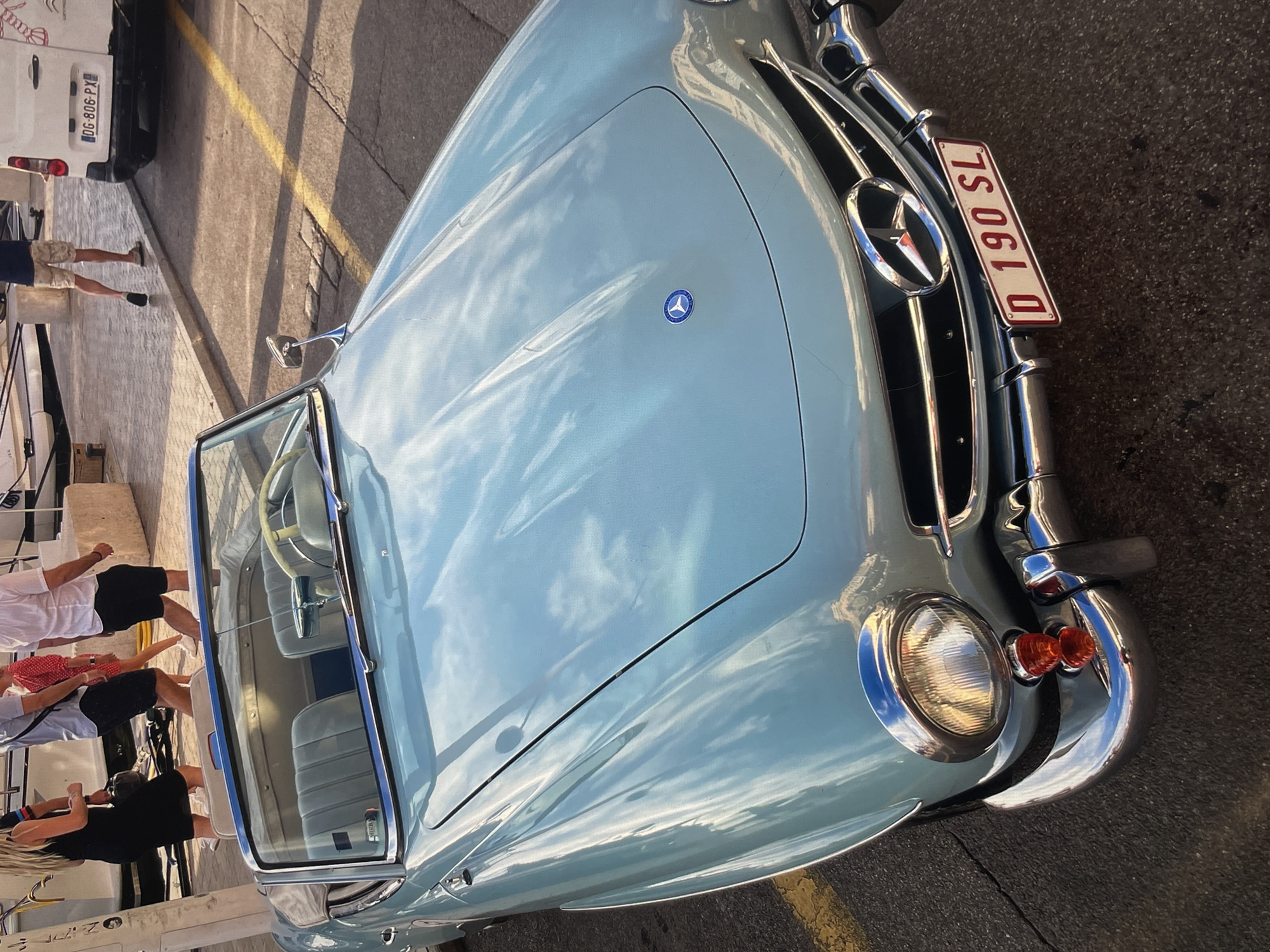 The image captures a vintage light blue Mercedes-Benz 190 SL convertible parked on a street with yellow lane markings. The car, the primary subject, is oriented with its front facing the viewer and slightly angled to the right. The front grille features the Mercedes-Benz logo, and a license plate reading "D 190 SL" is prominently displayed. The hood reflects the sky, indicating an outdoor location.
To the left of the car, several people are partially visible, suggesting a social gathering or event in proximity to the vehicle. They are engaged in possible conversations or observing the car.
The environment appears to be an urban setting, though the exact location is unspecified in the query. Details such as the road surface and the presence of other vehicles (a white van is visible in the background) corroborate this interpretation. The lighting suggests it could be late afternoon or early morning, given the shadows and the apparent warmth of the light.