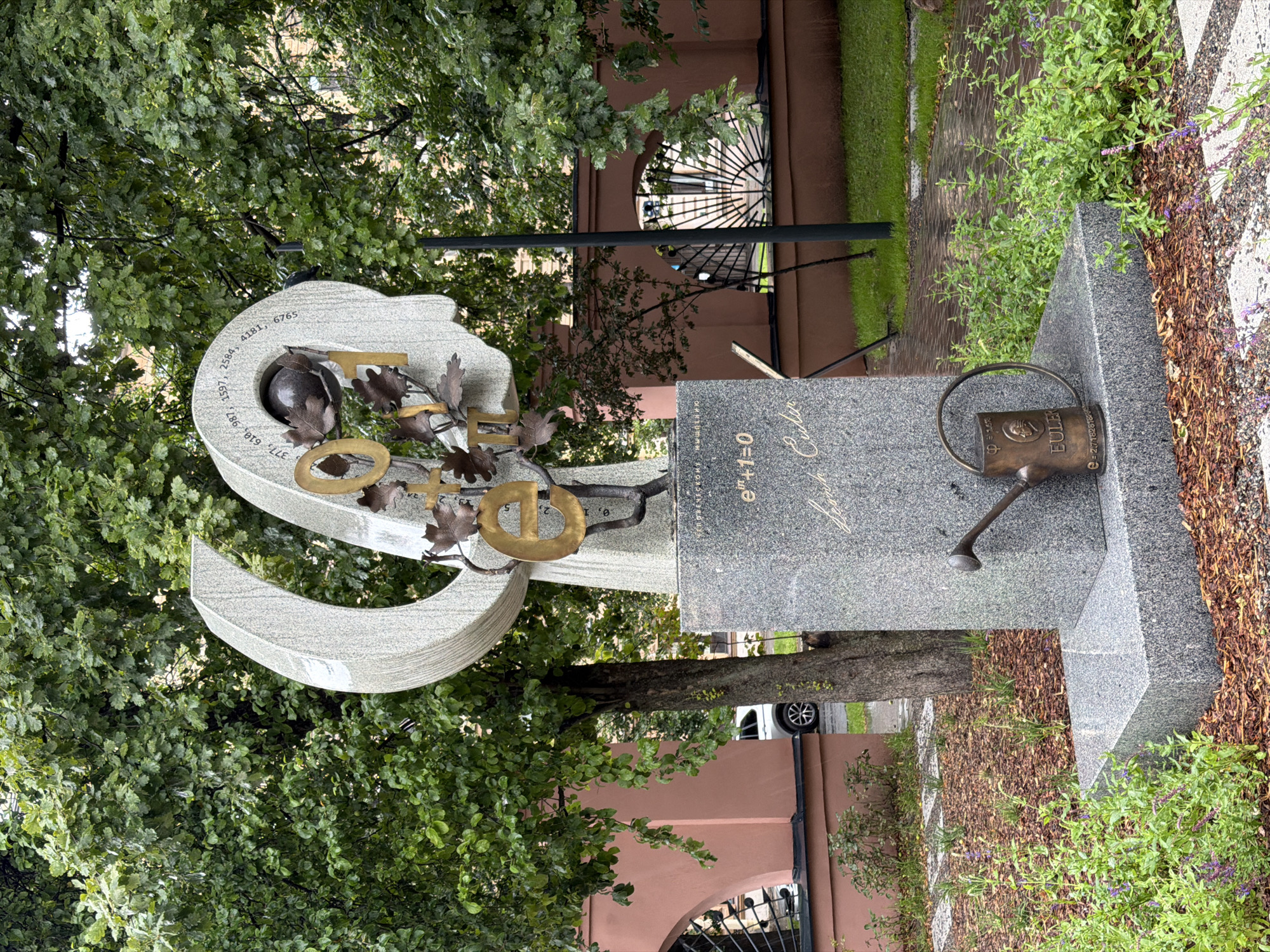This is a medium shot, outdoor, eye-level photograph of a monument dedicated to mathematician Leonhard Euler. The monument is located in the yard of the Saint Petersburg Mathematical Institute of the Russian Academy of Sciences, in Saint Petersburg, Russia.
The monument is made of granite and features a large, abstract sculpture resembling a human head with a crescent moon shape on the left. The head is formed by two curved stone elements, and inside one of the openings, there is a dark, spherical object, possibly representing a globe or an eye. Gold-colored letters and numbers, including "e", "π", and "1", are attached to the sculpture. The base of the monument is a tiered granite pedestal. On the front of the pedestal, the equation "e^(iπ) + 1 = 0" is inscribed, followed by what appears to be a signature or inscription in cursive. A bronze watering can with an inscription on its side reading "EULER" is placed in front of the pedestal.
The monument is surrounded by a landscaped area with mulch, grass, and small plants, some with purple flowers. In the background, there is a tree with green foliage, a reddish building with arched windows and barred gates, and a white car parked on a wet paved area. The overall atmosphere suggests a slightly overcast or rainy day due to the wet pavement.