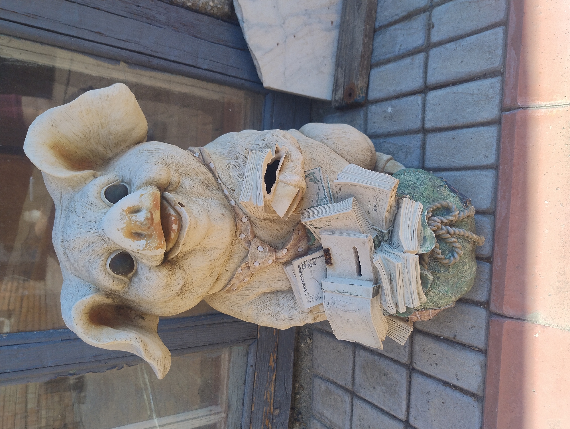 A weathered, light-colored statue of a pig sits on a paved walkway in front of a building with a glass window. The pig statue is dressed in a dotted tie and is holding stacks of money, some of which appear to be stylized representations of US dollars. The statue has a wide, open-mouthed smile and is positioned with its legs tucked underneath it, resting on a green, rope-like object. The background shows a wooden window frame and a glimpse of the building's interior through the glass. The paving stones are rectangular and arranged in a pattern.
