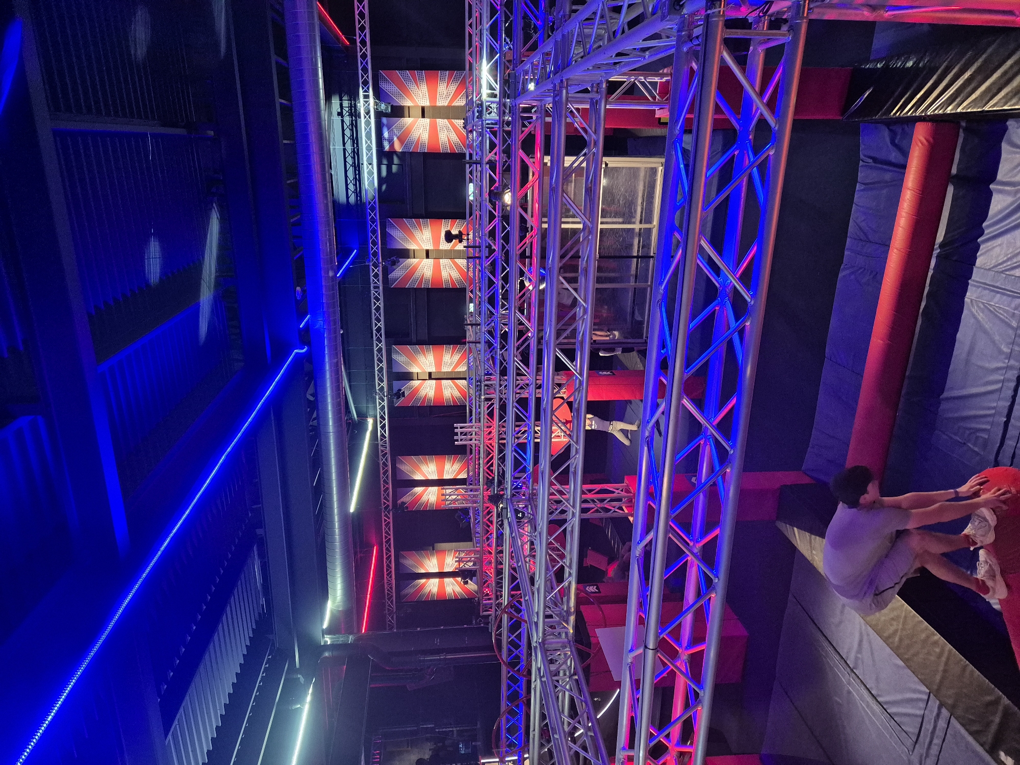 The image shows an indoor adventure park in Chelmsford, United Kingdom, featuring a ninja warrior course. The course is constructed with steel trusses illuminated in red and blue. Above, sections are decorated with the Union Jack flag. A young man is clinging to a padded obstacle, indicating participation in the course. The setting is a large indoor space with ventilation pipes running along the ceiling.