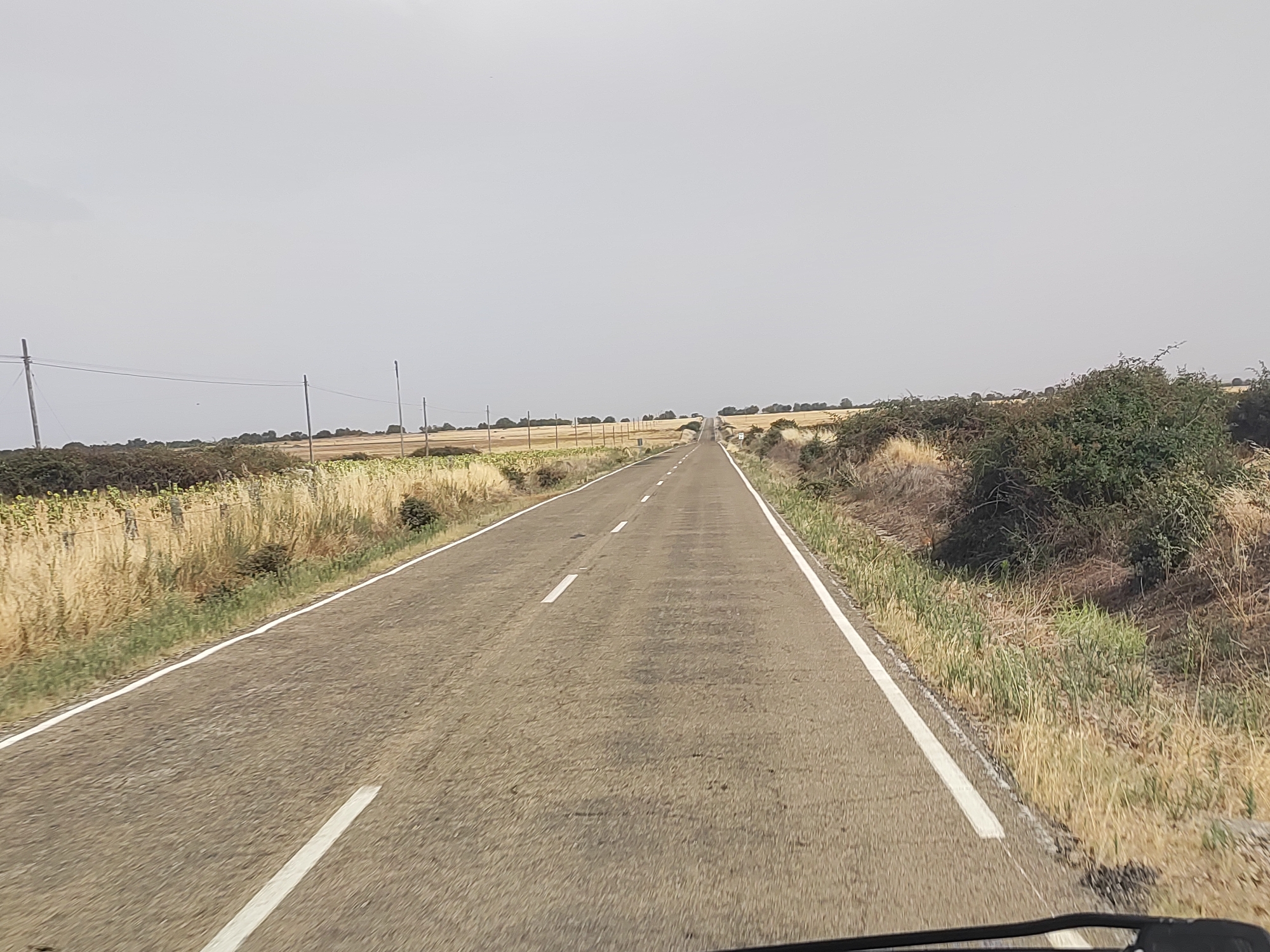 The image depicts a long, empty asphalt road stretching into the distance under a hazy, overcast sky. The road has a white line marking the edge on both sides and a broken white line down the center. The landscape flanking the road is dry and appears to be agricultural land with fields of tall, golden-brown grass or crops and scattered bushes and trees. There are several utility poles with wires visible along the left side of the road. The scene suggests a rural or semi-rural environment in Spain, possibly near Torrefrades. The perspective indicates the photo was taken from a vehicle moving forward. There are no people or animals visible in the frame. The overall impression is one of a quiet, possibly hot and dry, day in the countryside.