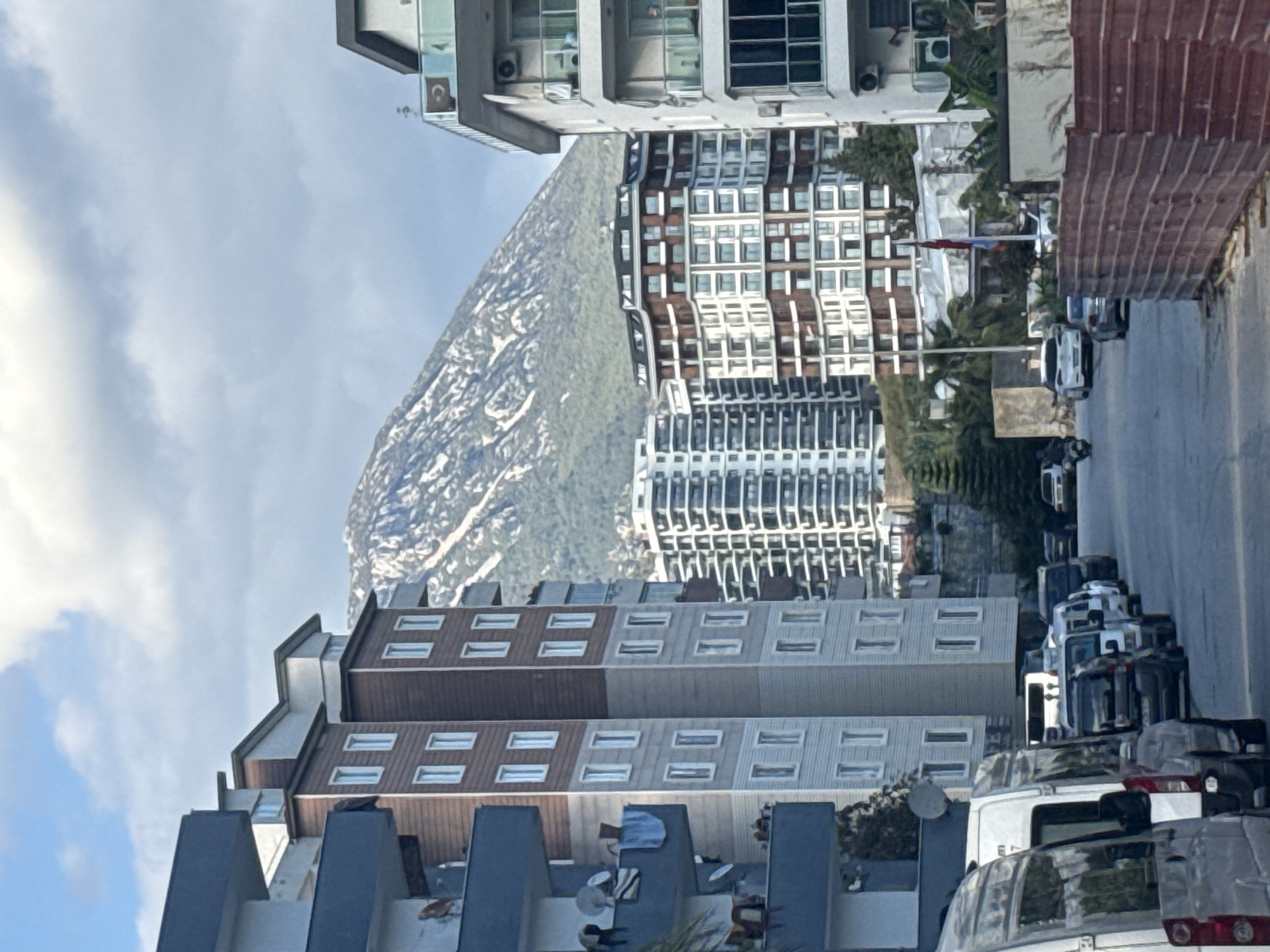 The image captures an urban scene with a prominent mountain backdrop. In the foreground, a modern building with a mix of brown and grey facades dominates the lower portion of the frame. A Turkish flag is visible amidst the buildings. A street is seen adjacent to the buildings, lined with parked vehicles. The backdrop is a majestic mountain, partially covered with snow. The sky above is a blend of blue and white, indicating daytime. The location is in the city of Unknown, Unknown.