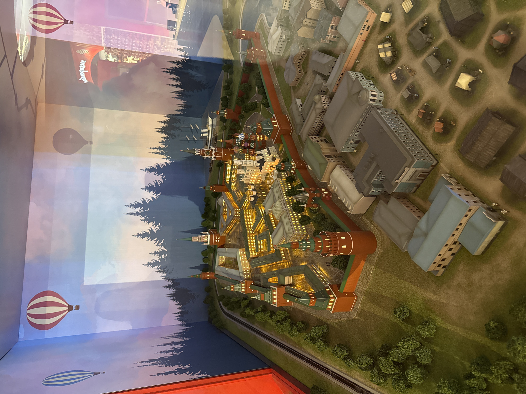 The image shows a miniature diorama of the Moscow Kremlin and adjacent areas. The Kremlin, with its distinctive red walls, towers, and cathedrals, is central in the model. Buildings are lighted from within. To the right of the Kremlin is a modern section with buildings and open spaces. In the background is a diorama of trees and a sky painted with hot air balloons. There is a glass enclosure creating reflections and the lighting is dim.
