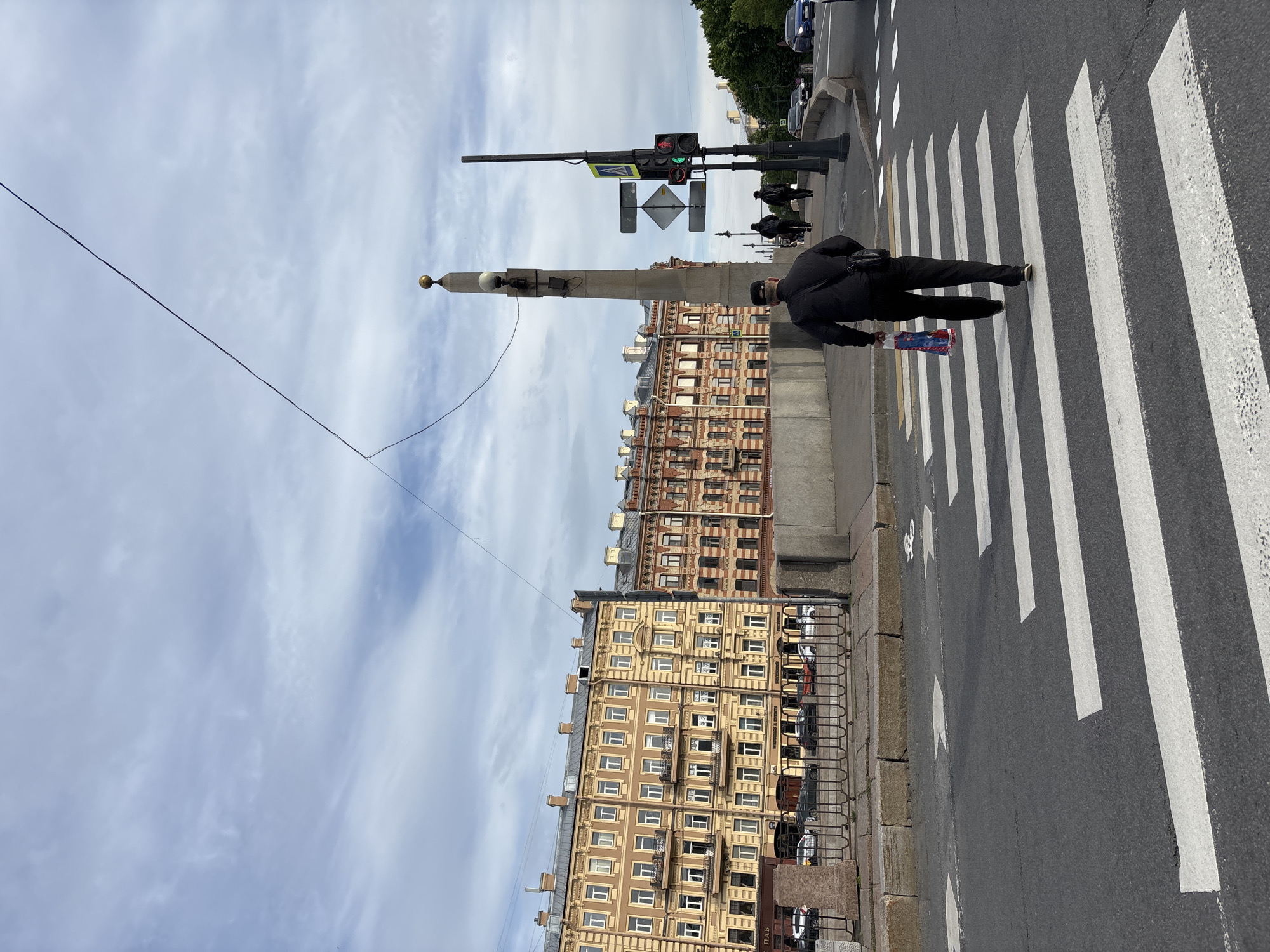 This media file captures an urban street scene in Saint Petersburg, Russia, under a bright, partly cloudy daytime sky. The image is dominated by a pedestrian crossing a street at a clearly marked zebra crossing. The person, viewed from behind, wears dark clothing and carries a brightly colored package in their left hand, which appears to have red, white, and blue elements and possibly the Cyrillic word "МИР" (MIR).
To the left of the pedestrian, a tall, light-colored street pole stands prominently, adorned with a golden finial at its top and equipped with a light fixture and other mounted devices. Further left, a complex traffic light system is visible, attached to a black pole. It includes a main vertical traffic light which shows a green signal, presumably for vehicular traffic. A separate, smaller pedestrian signal on an arm extending from the pole displays a green "walk" symbol, indicating that pedestrians are permitted to cross. Various street signs are also mounted on this pole, including a yellow diamond-shaped "Pedestrian Crossing" sign and white rectangular signs with black arrows.
Along the far side of the street, a row of multi-story buildings lines the road. The closest building features a warm, light brown or beige facade with numerous windows and balconies, showcasing classic European architectural elements. Further down the street, a building with red brick and lighter detailing is visible. A metal railing runs along the edge of the sidewalk, and parked cars are visible further down the street. Partially visible Cyrillic text, "ПАБ" (PAB), can be seen on a dark sign attached to the railing in the lower right corner. The asphalt road surface shows clear white stripes of the zebra crossing and some faded white directional arrows.