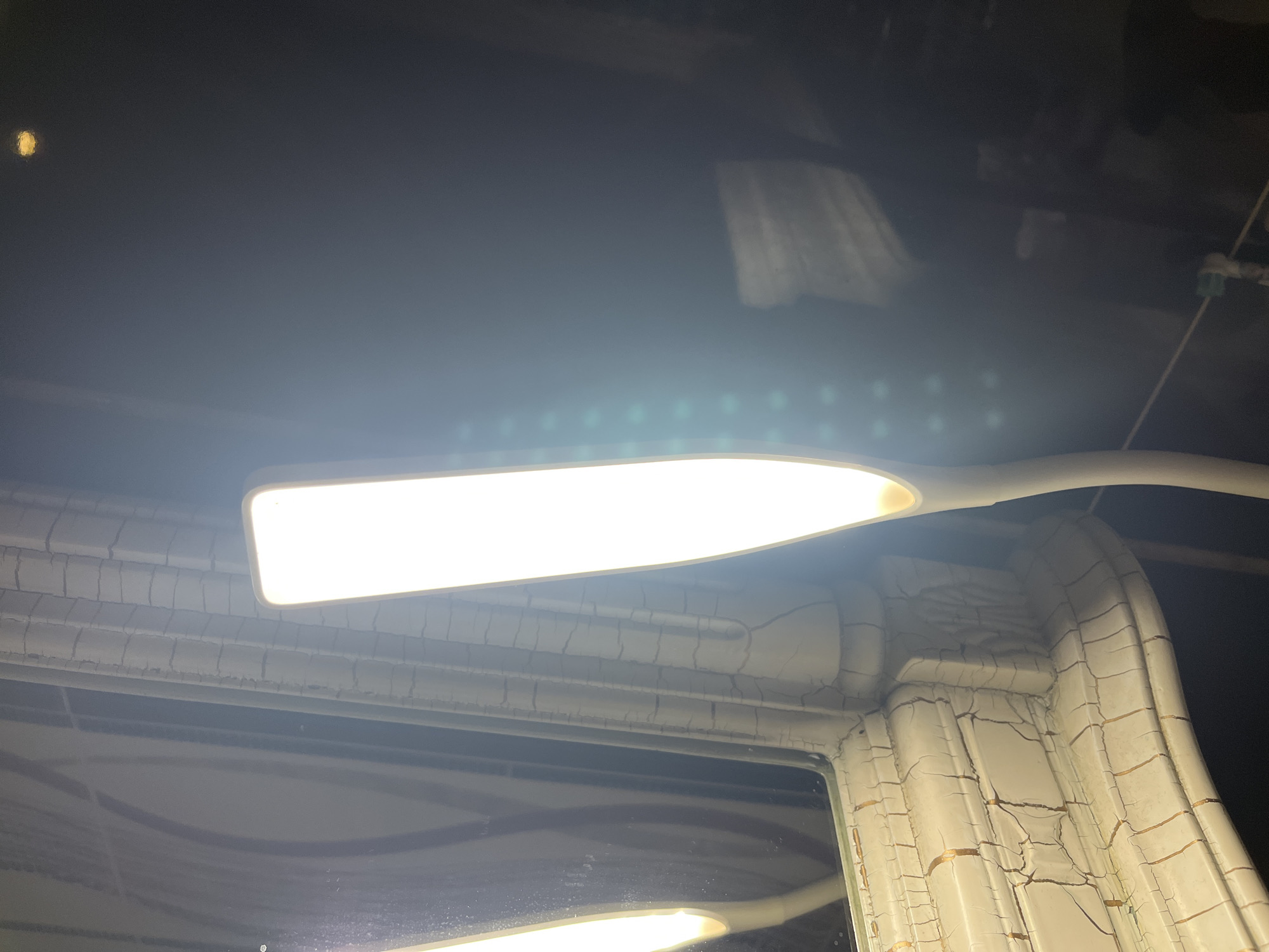 A close-up, eye-level shot shows a bright white LED lamp with a flexible gooseneck stem extending from the right side of the frame. The lamp is illuminated and positioned in front of a dark background with a slight sheen, possibly a wall or a cabinet. To the left of the lamp, a decorative mirror frame is visible. The frame has an antique or distressed appearance with a crackled white finish and thin gold lines accentuating the crackle pattern. The mirror itself reflects the lamp and a textured, vertical surface, possibly curtains or a wallpaper pattern, which appears to be white with gray lines. The setting is an indoor environment, potentially a bedroom or a dressing area. The location is identified as Tarnivtsi, Ukraine, though there are no visual elements in the image that specifically confirm this location.