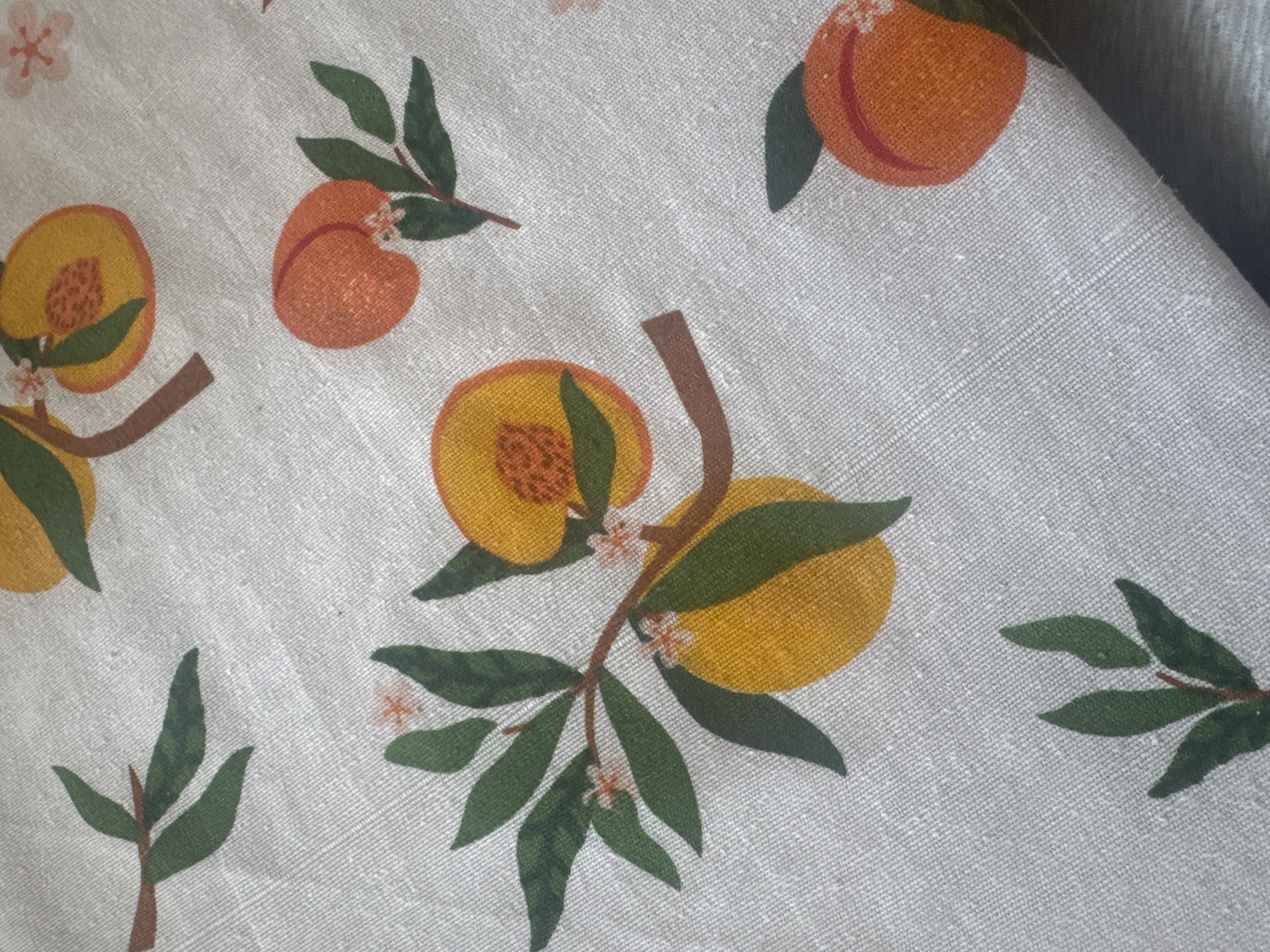 The image shows a piece of light gray fabric with a repeating pattern of peaches, leaves, branches, and small flowers. The peaches are depicted in shades of yellow and orange, with visible details like the peach fuzz and pit. The leaves are green, and the branches are brown. The pattern is scattered across the fabric. There is no indication of the location being Sofia, Bulgaria.