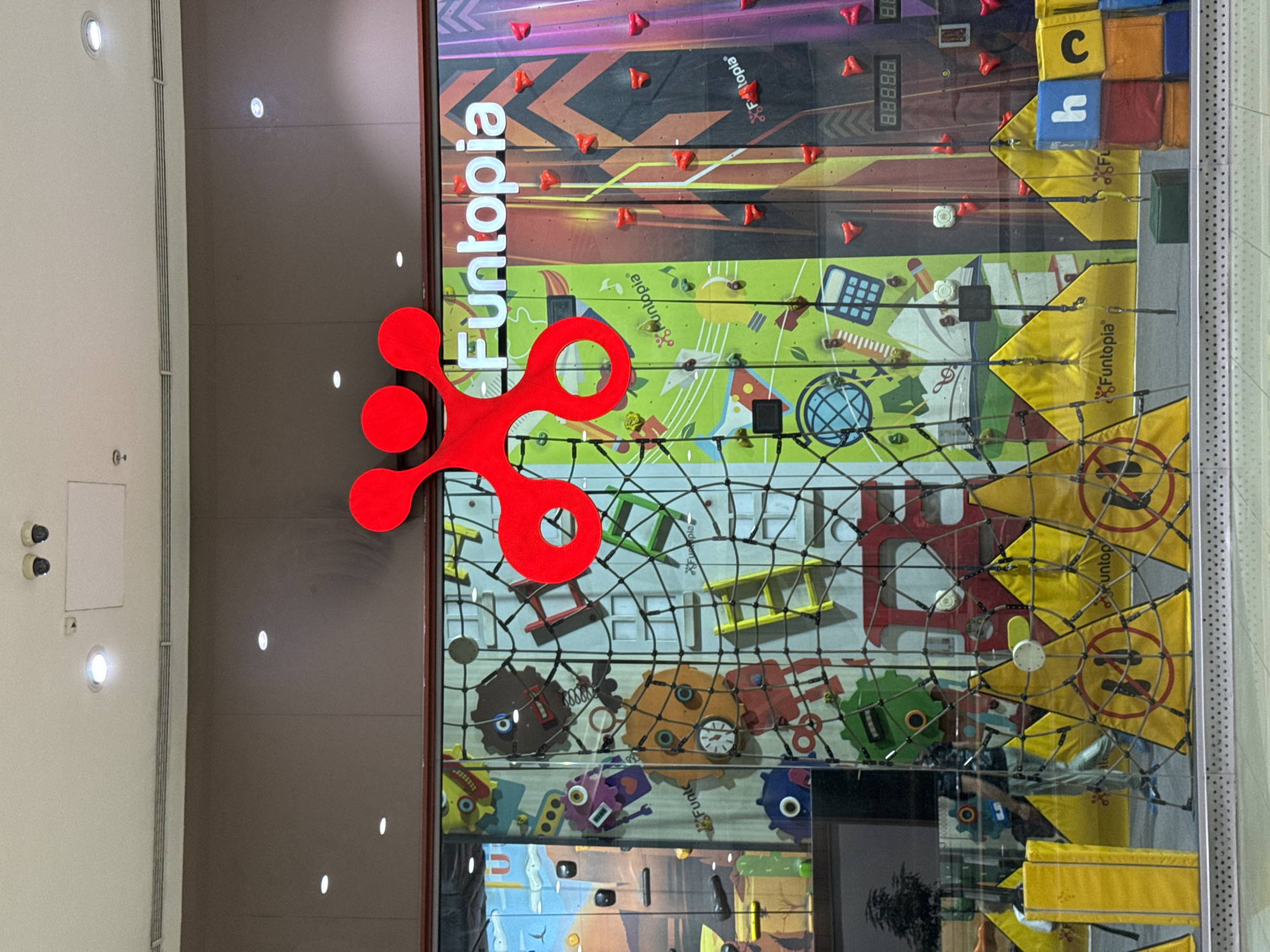 This image displays a vibrant indoor climbing and play facility, "Funtopia", located in Sofia, Bulgaria. The central focus is a tall, multi-paneled climbing wall designed for recreational activity. The wall features diverse, colorful patterns including gears, cartoon robots, a calculator, a globe, and numerical sequences like "88888". Numerous red climbing holds are scattered across these panels.
A black safety net is visible over sections of the climbing wall, with yellow padded elements behind it. Some of these yellow pads display a red "no entry" or "no stepping" symbol and small "Funtopia" brand names. A prominent, large red, multi-lobed "Funtopia" logo is positioned on the left side of the wall, with the brand name "Funtopia" written vertically in white text next to it.
To the left of the climbing structure, the interior of a modern building is visible, characterized by light-colored walls with recessed lighting and two security cameras. The setting is brightly lit, suggesting an inviting and engaging environment for active play. Other visible text elements include the letters "C" and "H" on colorful block-like structures integrated into the wall design. No people are visible, but the setup clearly indicates an area intended for fun and adventure.