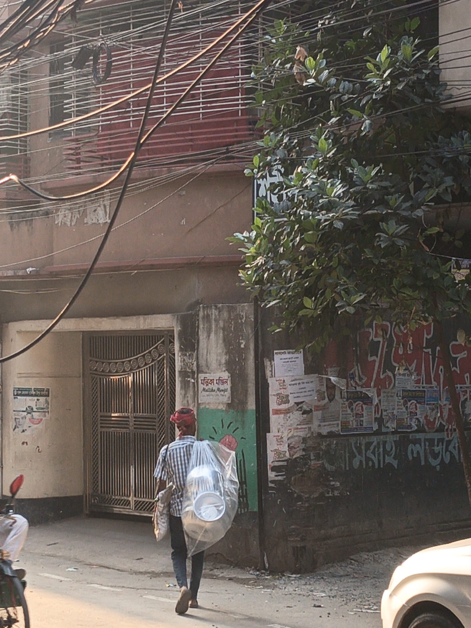 A man with a red head covering walks away from a building in Dhaka, Bangladesh. He carries a large, clear plastic bag filled with white objects, possibly dishes, over his right shoulder. He wears a checkered shirt, dark pants, and sandals.
The building is two stories high with an iron-barred gate. A sign is visible next to the gate. Posters are plastered on the wall beside a tree on the right. Power lines crisscross above the building. A motorcycle and part of a car are visible at the edges of the frame. The area is a street with weathered buildings.