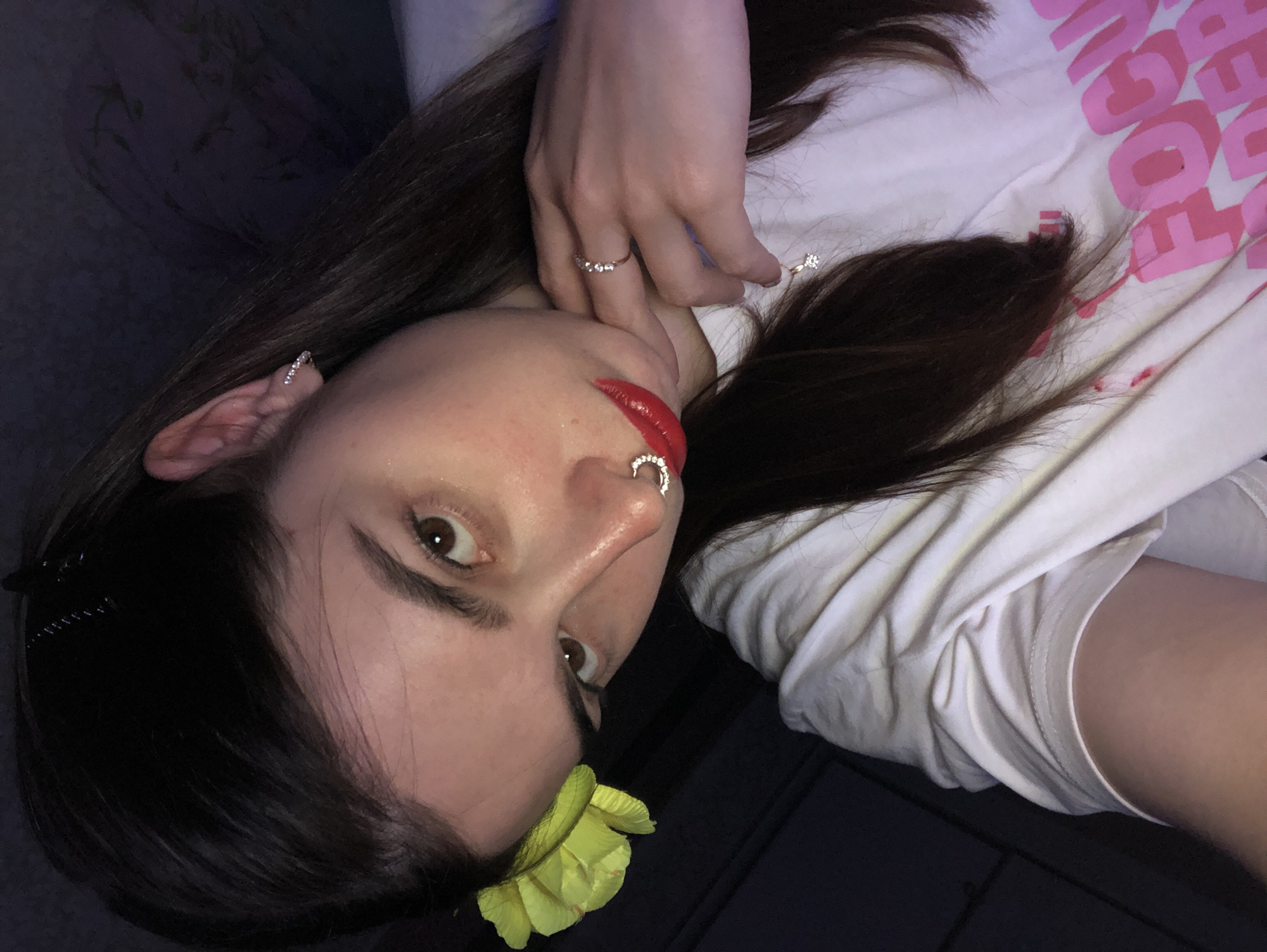 A person with dark hair, red lipstick, and multiple piercings (ear, nose) is reclining. The person is wearing rings on their fingers and a t-shirt with pink lettering. A yellow flower is near their jawline. The background is dark, suggesting an indoor setting. No information is visible to suggest this is in the city of Noviy, Russia.