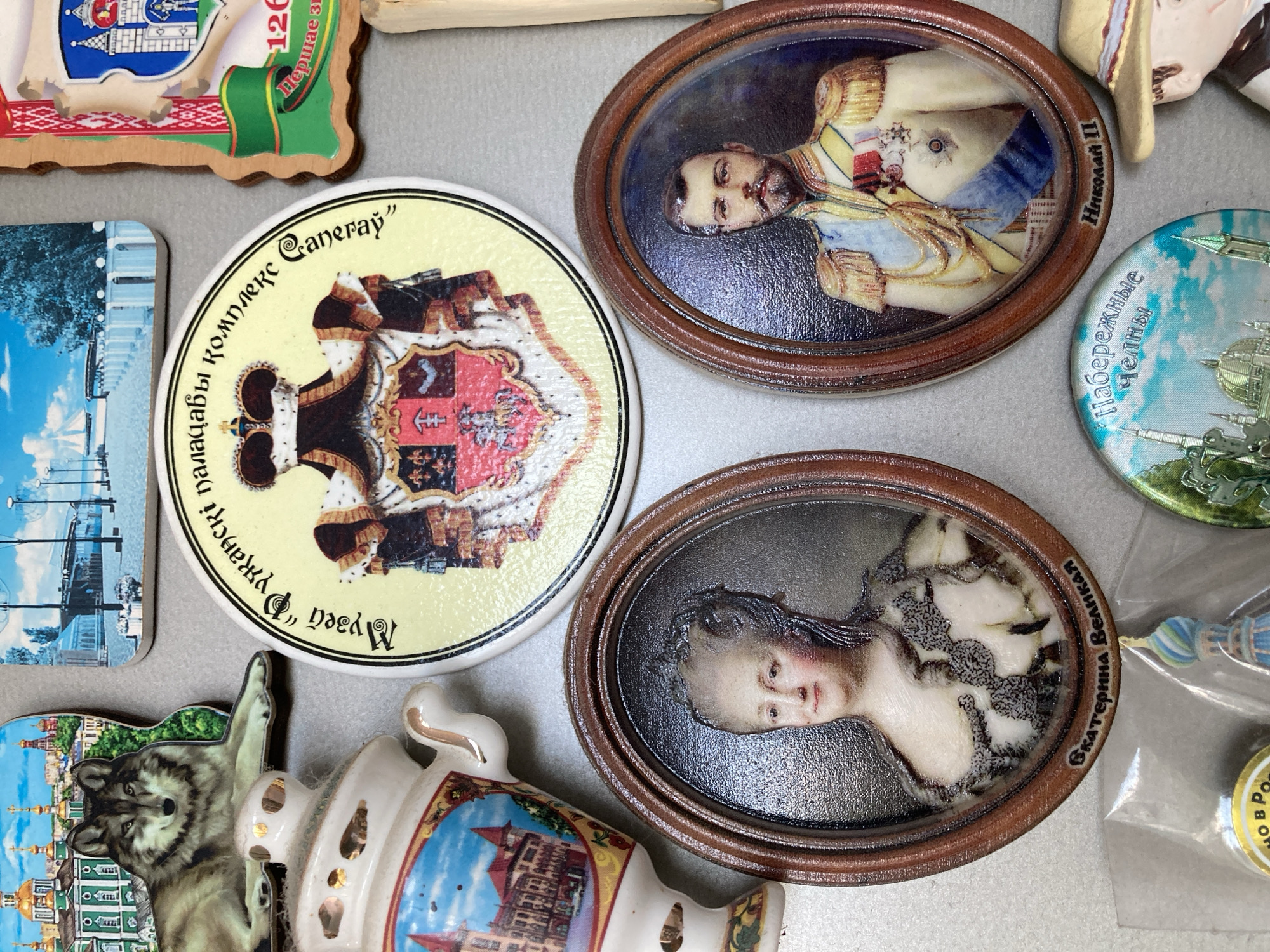 The image shows a collection of decorative magnets. In the upper left, a magnet depicts a wolf and a samovar with buildings. To the right of that, a round magnet features a royal coat of arms and text in Cyrillic script. A wooden magnet with a shield and text is in the upper right corner. Below these, two oval magnets display portraits: one of a woman and the other of a man in military attire. Text below the portraits identifies them as "Екатерина Великая" (Catherine the Great) and "Николай II" (Nicholas II). In the lower section, a round magnet shows buildings and text, and a straw hat sits on a bust of a person. The background is a neutral gray surface.