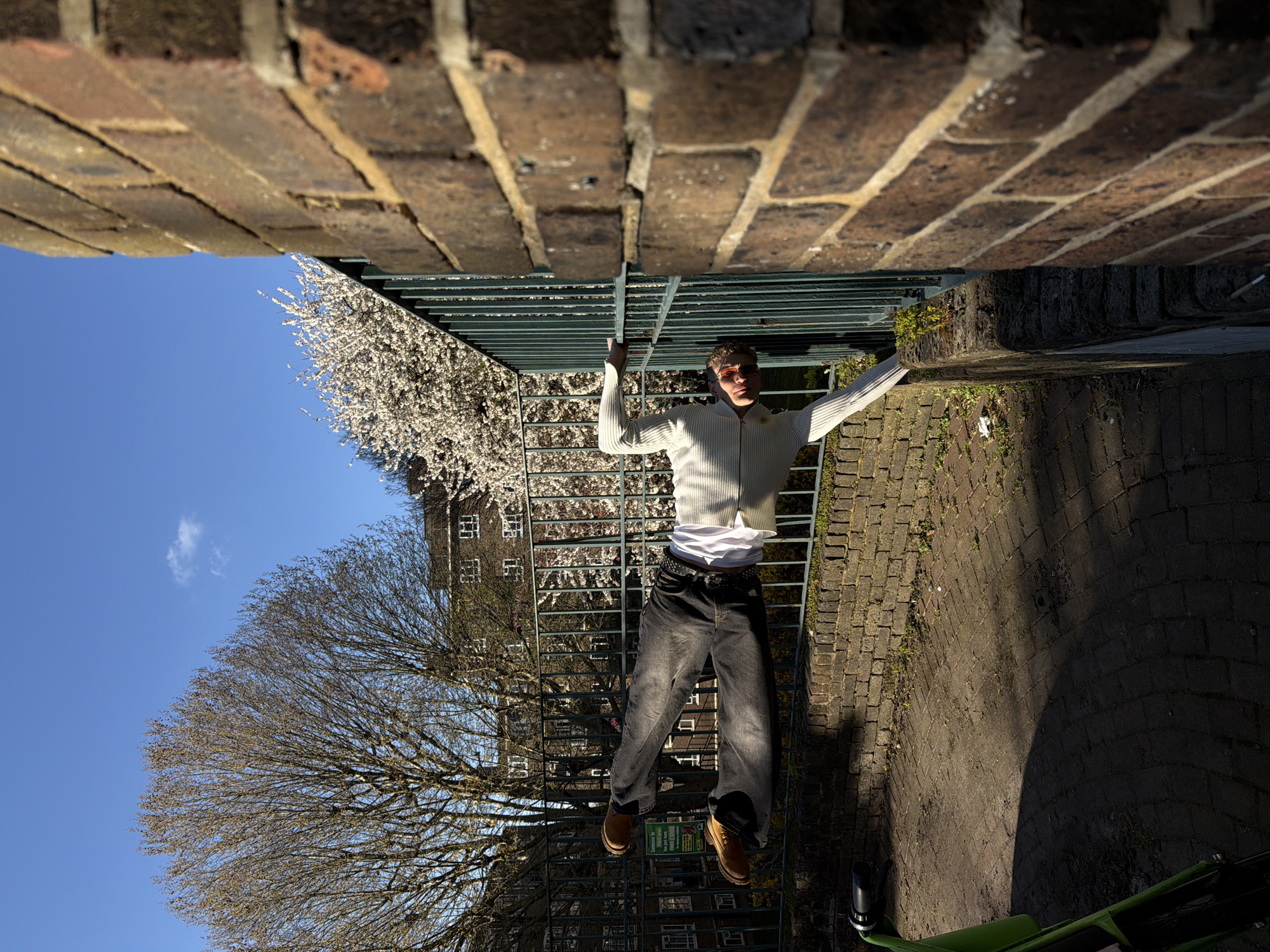 A person in a white sweater, dark jeans, and brown boots is hanging from a metal railing. The person is wearing sunglasses and a white t-shirt under the sweater. The railing is between two brick structures. Behind the railing is a tree with bare branches on the left and a flowering tree on the right. A building with windows is visible behind the trees. Below the person, there is a brick paved area with a green bicycle. The sky is blue with a small cloud. The image is taken from a low angle and is rotated. This appears to be an urban environment. The exact location in the city of London, United Kingdom cannot be identified from the provided image.