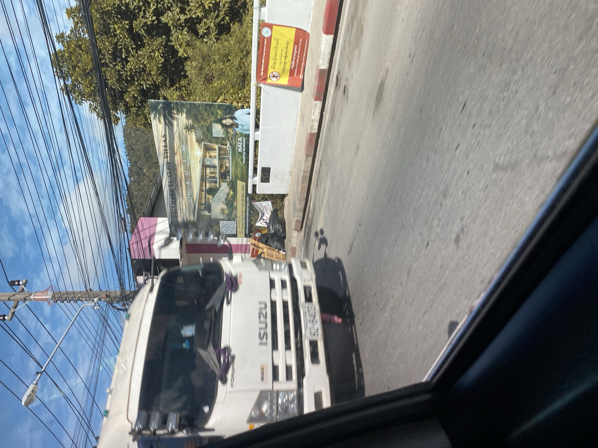 In Baan Hua Thanon, Thailand, a white Isuzu truck is captured in motion, viewed from the perspective of a passenger inside another vehicle. The truck's front is clearly visible, showing its headlights, grille, and the "ISUZU" logo. A license plate with Thai characters and numbers is affixed to the front bumper. The truck has purple decorative tassels hanging from the windshield.
Beyond the truck, a billboard advertises "PRIVILEGE LUXURY VILLAS" with an image of a modern house and a woman. To the right of the billboard, a white concrete barrier runs along the roadside, adorned with two banners. One banner, red and yellow, features text and a symbol of a prohibition sign. The other banner, white and smaller, also displays text and a similar prohibition symbol. Black garbage bags and wooden planks are piled near the base of the barrier.
The environment is characterized by a clear blue sky with scattered clouds, overhead electrical wires strung across wooden poles, and lush green trees in the background. The road surface is asphalt, with painted red and white stripes marking the curb. The image conveys a sense of everyday life in a bustling Thai town, with commercial advertising and traffic contributing to the scene.