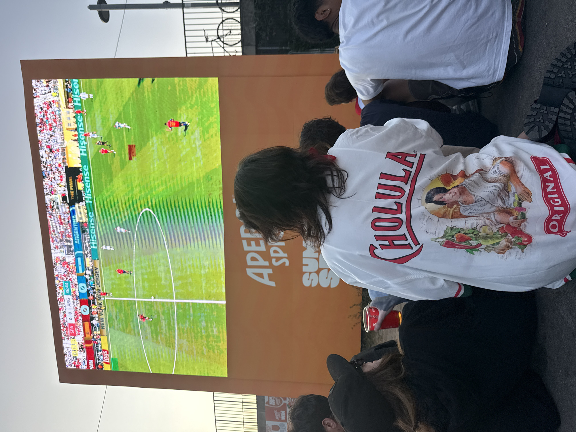 In an outdoor urban setting, a group of people are gathered to watch a large screen displaying a soccer match. The screen shows a game between Spain and Argentina, with the score at 0-0 in the 36th minute. The crowd is seated on the ground, facing the screen. One person in the foreground is wearing a white shirt with a graphic design featuring a woman and vegetables, with the word "CHOLULA" and "ORIGINAL" prominently displayed. Another person next to them is holding a red cup. In the background, a bicycle is visible in front of a railing. The overall atmosphere appears to be one of relaxed engagement with the sporting event.