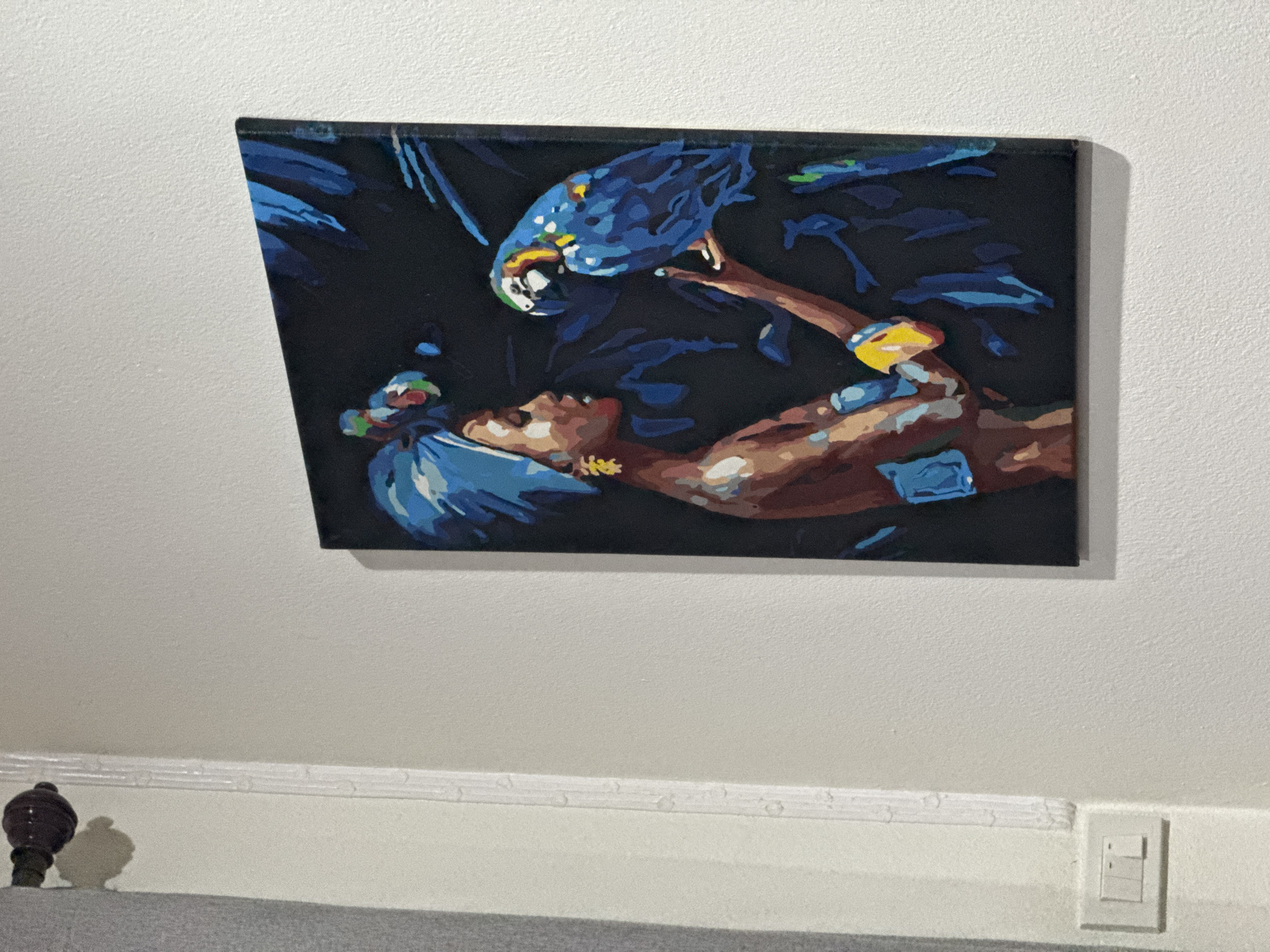 A painting of a woman in a blue headwrap and a blue parrot on her hand is hanging on a textured white wall. The painting depicts the woman in profile, facing left, with her head tilted down towards the parrot. The parrot is mostly blue with some yellow and green markings. The background of the painting is black with abstract blue shapes. To the left of the painting, there is a dark brown curtain rod with a decorative knob and a white molding that runs vertically down the wall. Below the molding, on the lower left side of the wall, there is a white double light switch. The scene appears to be indoors, as suggested by the presence of the light switch and the painting on the wall.