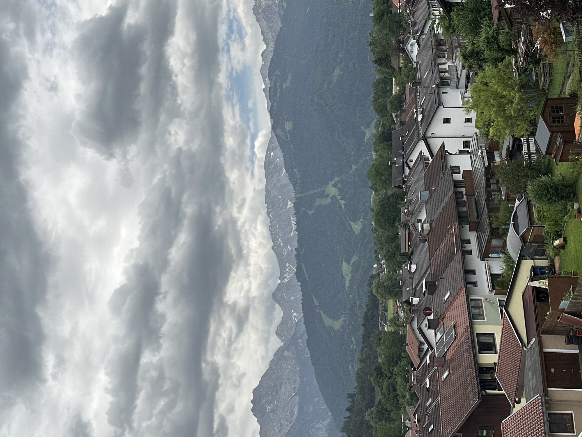 This is an aerial, full-frame, eye-level, medium shot of a residential area in Garmisch-Partenkirchen, Germany, under a cloudy sky. In the foreground, there are several houses with reddish-brown tiled roofs, some with solar panels. There are also some sheds, fences, and greenery, including trees and grass. In the middle ground, there are more houses nestled among trees. In the background, a dense forest covers rolling hills, and behind that, majestic, snow-capped mountains rise under the dramatic clouds. A ski slope is visible on one of the mountains. The scene appears to be a quiet, suburban or semi-rural setting. There are no people or animals visible in the image. The overall mood is overcast and slightly muted due to the cloudy weather.