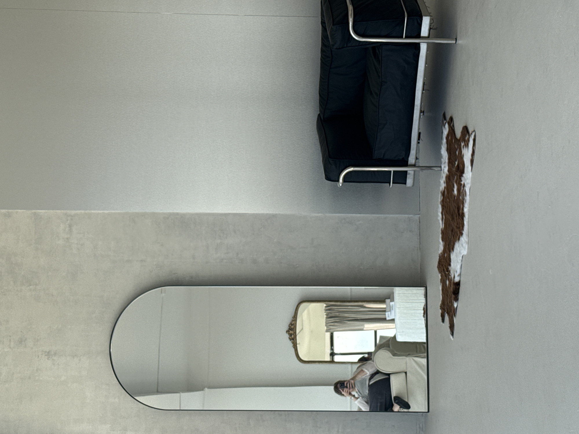 The image depicts an interior scene with a modern aesthetic. Dominating the upper right quadrant is a black leather sofa with a chrome frame. Adjacent to the sofa, a brown and white animal hide or decorative rug hangs vertically on the wall.
In the lower left quadrant, a large, rounded-rectangular mirror reflects a portion of the room. Inside the reflection, a person holding a phone is visible, capturing a scene with a second, smaller ornate gold-framed mirror, a white sofa, and a window. The reflection suggests the room has a light and airy atmosphere.
The walls are a uniform gray, providing a neutral backdrop for the furnishings. The overall impression is a clean, minimalist space with a touch of character introduced by the rug and the varied mirror styles. There is no information to ascertain the location of Izhevsk, Russia.