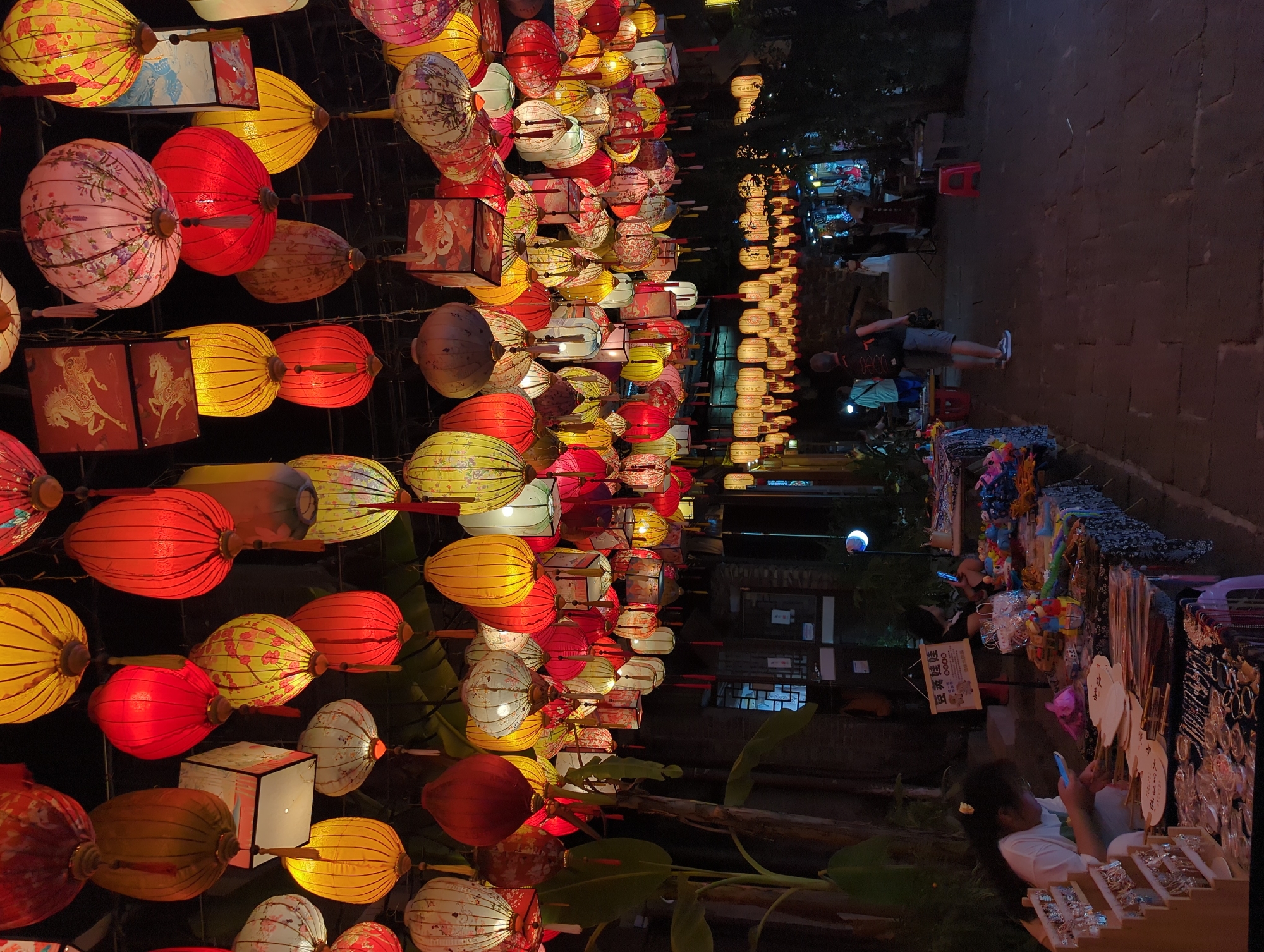 The image depicts a night scene in Fenghuang, China, a historic town known for its well-preserved traditional architecture and vibrant cultural atmosphere, particularly at night when the lanterns are lit. The scene is dominated by a multitude of colorful lanterns hanging overhead, creating a festive and inviting ambiance. These lanterns are of various shapes, sizes, and designs, with some featuring intricate patterns and others solid colors.
In the foreground, a market stall is set up with various goods displayed on tables. A young girl, likely a vendor or helper, is seated at the stall, looking at her mobile phone. She is wearing a white t-shirt. Several other people are visible in the background, some browsing at stalls and others walking through the area. The ground is paved with cobblestones, characteristic of many ancient Chinese towns. The architecture visible in the background appears to be traditional, with dark wood and tiled roofs, further enhancing the historical feel of the location. The overall impression is one of a lively night market in a picturesque, traditional setting.