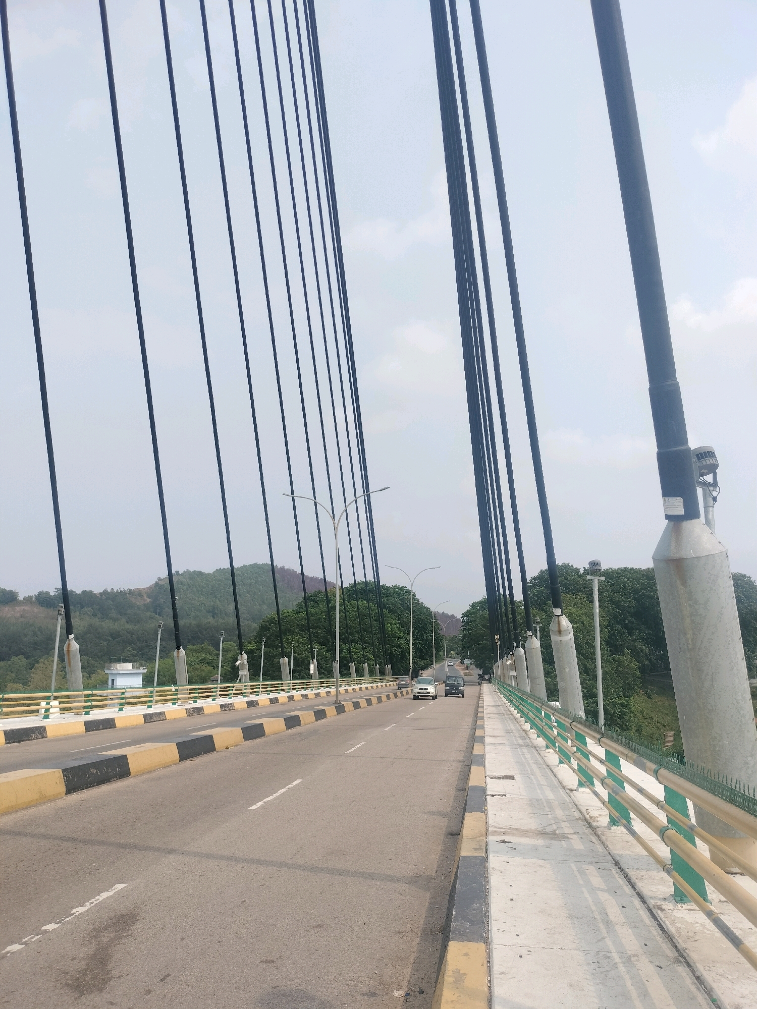 The image shows a view from a suspension bridge, likely the Batam-Bintan bridge in Batam, Indonesia, looking across the road. The bridge features numerous dark, vertical suspension cables extending from the top of the frame. The roadway, marked with white lines and a yellow and black striped curb, carries a few cars and vans. To the right of the road is a concrete pedestrian walkway with a yellow and green railing. In the background, lush green trees and rolling hills under a hazy sky suggest a natural, semi-urban or rural setting. The sky is overcast with a light grey hue.