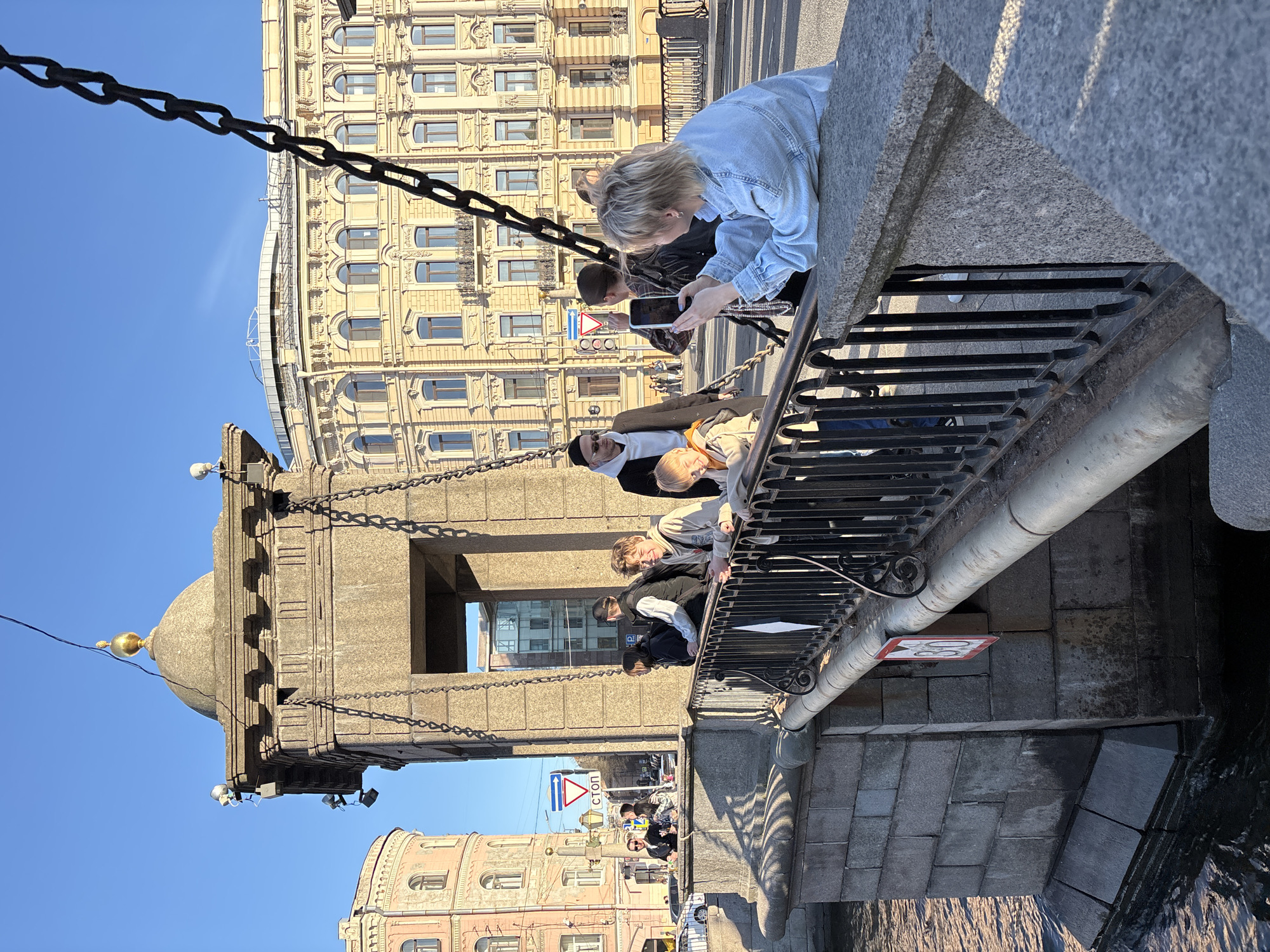 The image shows a group of people leaning over the railing of the Palace Bridge (Дворцовый мост) in Saint Petersburg, Russia. A woman in a denim jacket is holding a smartphone and appears to be taking a picture. Several other people are also looking over the railing. In the background is the Winter Palace and other buildings along the Neva River. The bridge's chain mechanism and decorative tower are visible. The sky is clear and blue.