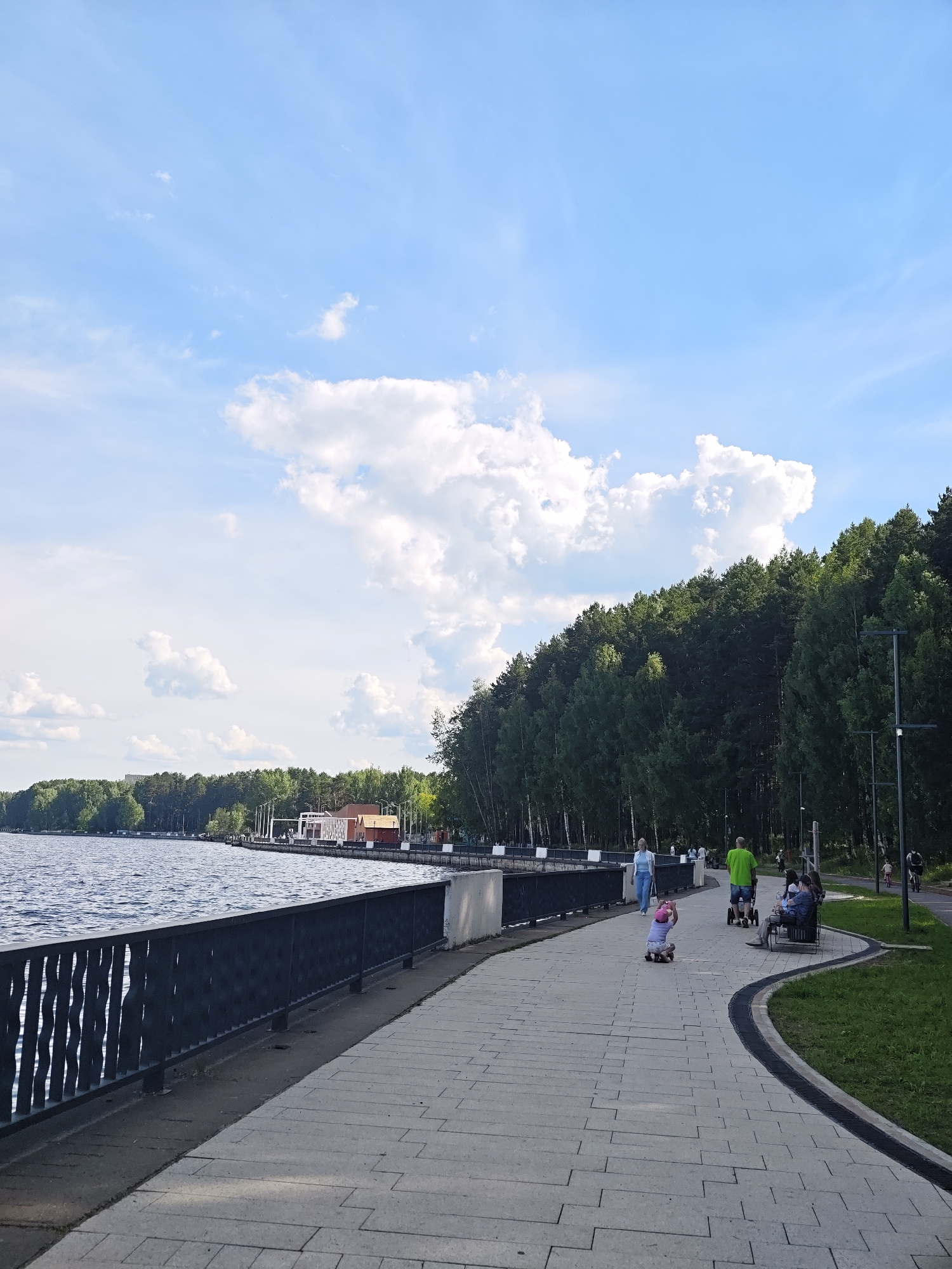 This image depicts a scenic waterfront promenade on a bright, partly cloudy day. The scene is set in Chaikovsky, Russia, and the time of day appears to be afternoon, given the bright sunlight and the shadows cast.
The foreground features a wide, paved walkway made of light-colored rectangular stones arranged in a geometric pattern. A dark, decorative metal fence runs along the left side of the walkway, separating it from a calm body of water. The water is a deep blue-gray, reflecting the sky and appearing slightly rippled.
Along the promenade, several individuals are engaged in leisure activities. A young child in a pink helmet is on a small ride-on toy, moving across the path. Further along, a woman in a light blue top and jeans walks, while a man in a bright green t-shirt pushes a stroller. A group of people are seated on a bench, enjoying the view. In the background, a dense line of green trees forms a natural backdrop to the promenade, with a few buildings peeking through the foliage. The sky is a vibrant blue, dotted with fluffy white clouds.
The overall atmosphere is one of peaceful recreation and enjoyment of nature. The well-maintained walkway, the presence of families and individuals relaxing, and the natural beauty of the water and trees suggest a popular public space for outdoor activities. No visible text is present in the image.