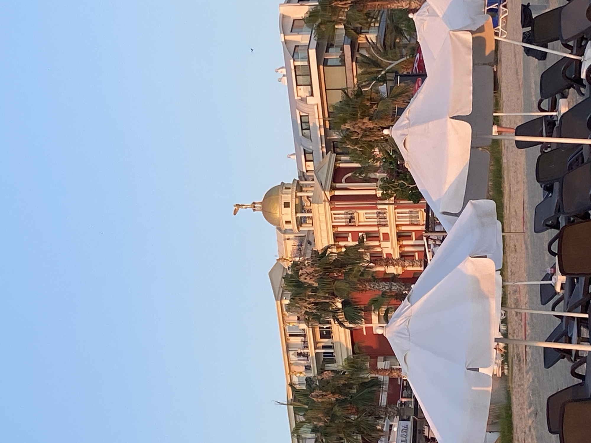 The image depicts a beach scene in Alanya, Turkey, with a large resort building in the background. Several white beach umbrellas are in the foreground, casting shadows on the sand. Beach chairs are visible beneath the umbrellas. The resort building features a distinctive golden dome topped with a statue and has multiple levels with balconies. Palm trees are interspersed throughout the scene. The sky is clear and blue, suggesting a sunny day.