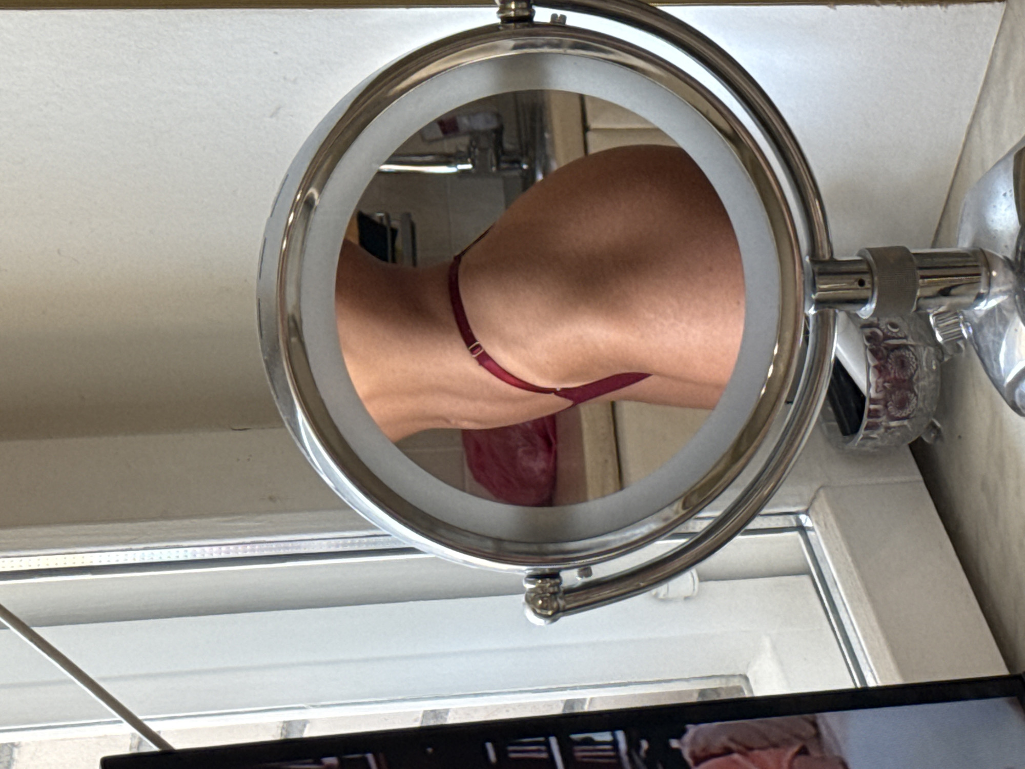 A mirror sits on a countertop in front of a window. The mirror reflects a person's side, specifically their hip and lower back, clad in red underwear. In the background of the reflection, a sink and cabinets are visible, suggesting a bathroom or kitchen setting. To the left of the mirror, a portion of a laptop screen is visible, displaying an image of two people. The window behind the mirror looks out onto a building with a brick facade. The overall scene appears to be indoors, possibly in an apartment. There are no specific landmarks that definitively place this scene within Saint Petersburg, Russia.