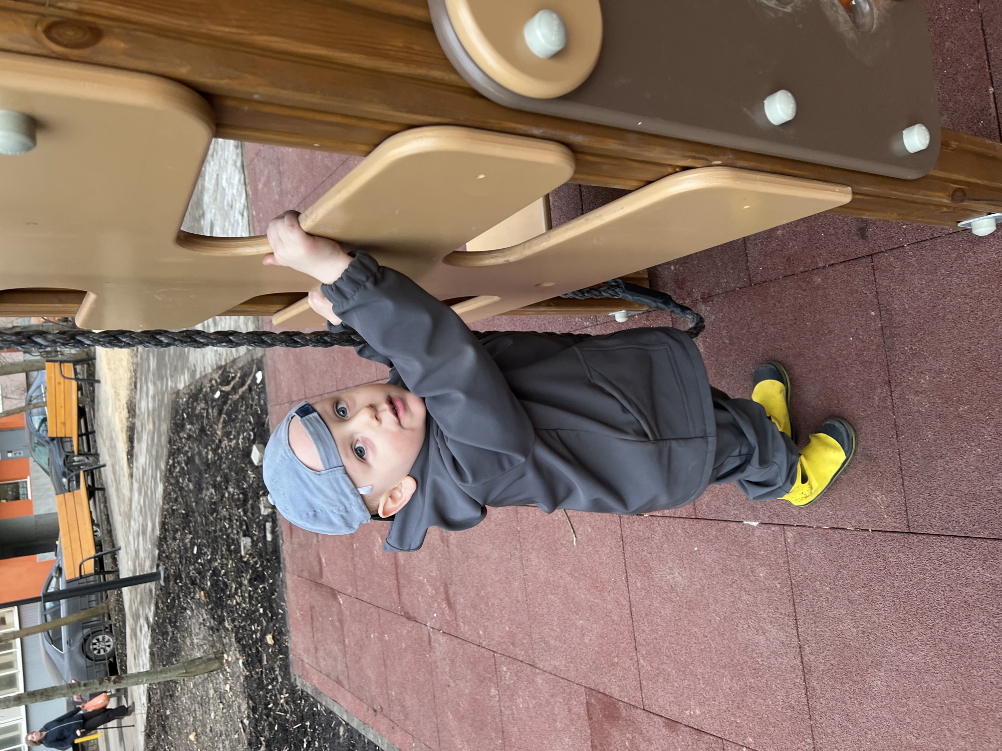 A young child in a grey hooded jacket, grey hat, and yellow boots is hanging upside down from a wooden playground structure in Mytishchi, Russia. The structure has brown wooden rungs. The ground is covered in red rubber tiles. In the background, there is a parking lot with cars, trees, and buildings, one with an orange facade. A person wearing a dark jacket and yellow pants or possibly carrying a yellow item is seen walking in the background.