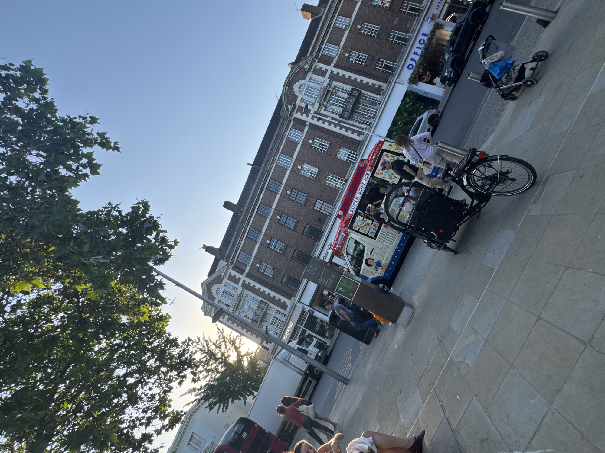 The image depicts a street scene in an urban area. A brick building with multiple windows and what appears to be an "OFFICE" sign is prominent in the background. A commercial ice cream truck is parked along the sidewalk. People are present, including individuals near the ice cream truck and others walking on the sidewalk. A bicycle-style vehicle with a carrier is parked next to the truck. The sky is visible in the upper left corner, suggesting it is daytime. Street furniture and vehicles are also visible.