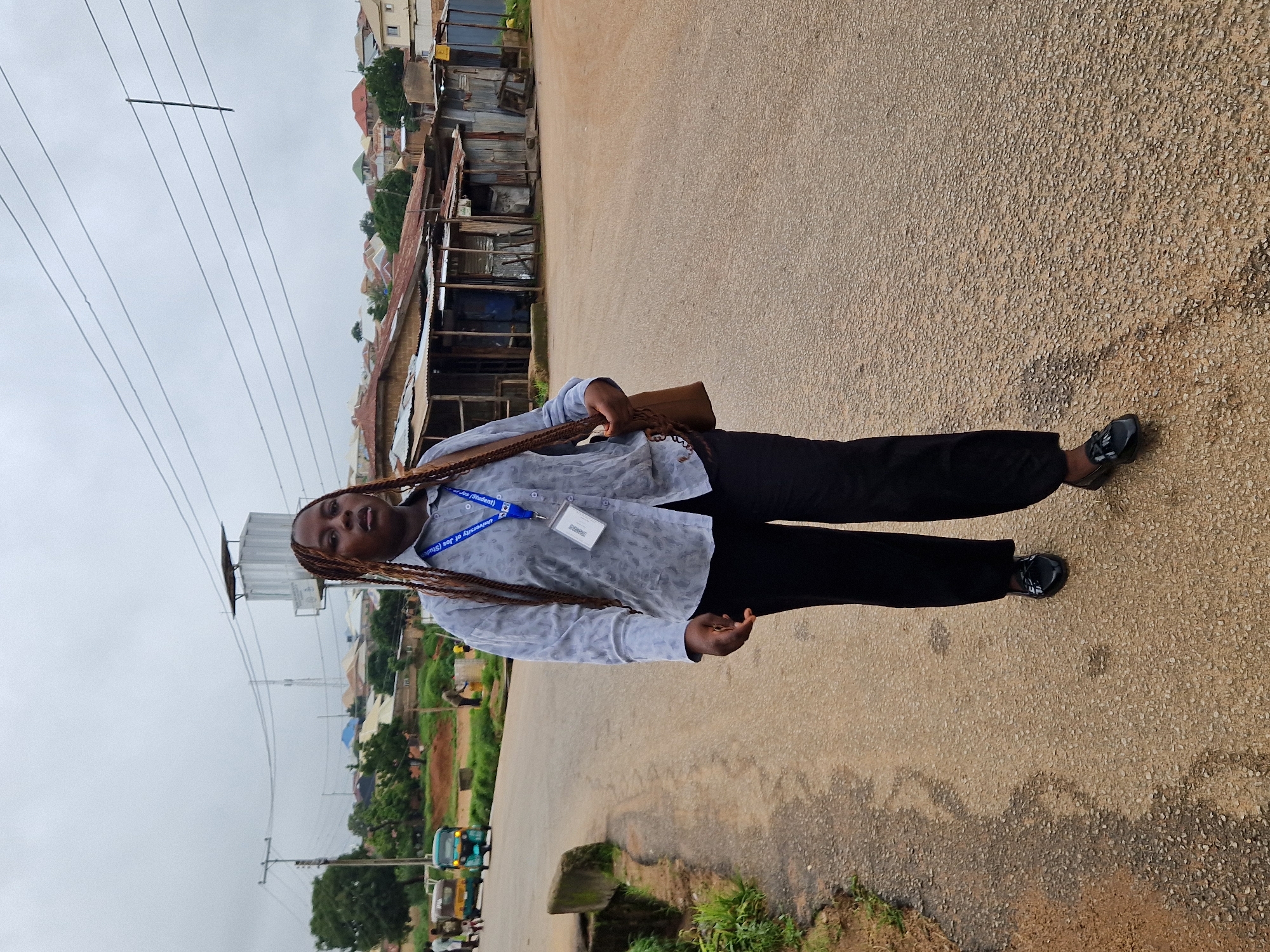 A young Black woman with braided hair walks on a paved road in what appears to be a semi-urban area, possibly in Kabong, Nigeria. She is wearing a light blue patterned long-sleeved shirt with a lanyard around her neck, black pants, and black slip-on shoes. A brown shoulder bag is slung over her left shoulder. The road is flanked by a grassy embankment on the left and a row of informal structures made of corrugated metal and wood on the right. In the background, there are more buildings, trees, and utility poles, suggesting a residential or commercial area. The sky is overcast. Several three-wheeled vehicles are visible in the distance. The overall scene depicts a daily life moment in a developing urban environment.