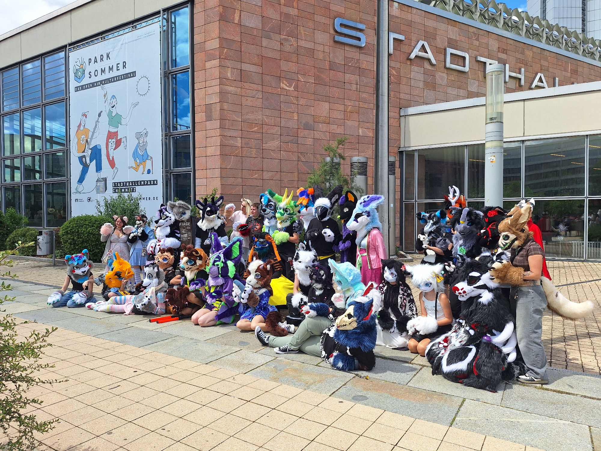A large group of people, many of whom are wearing elaborate furry costumes resembling animals, are gathered outside a modern building with a brick facade. The building has large glass windows and a prominent sign that reads "STADTHALLE" in large silver letters. Above the entrance, a banner advertises "PARK SOMMER DAS OPEN AIR KULTURFESTIVAL" and "STADTHALLENPARK CHEMNITZ". The gathering appears to be a public event, likely a festival or convention, given the costumes and the outdoor setting. The ground is paved with light-colored square tiles. Some individuals in the group are sitting on the ground, while others are standing. The weather is overcast, suggesting a temperate climate. The people in the furry costumes are diverse, with various animal species and colors represented, indicating a community gathering for enthusiasts of this particular subculture.